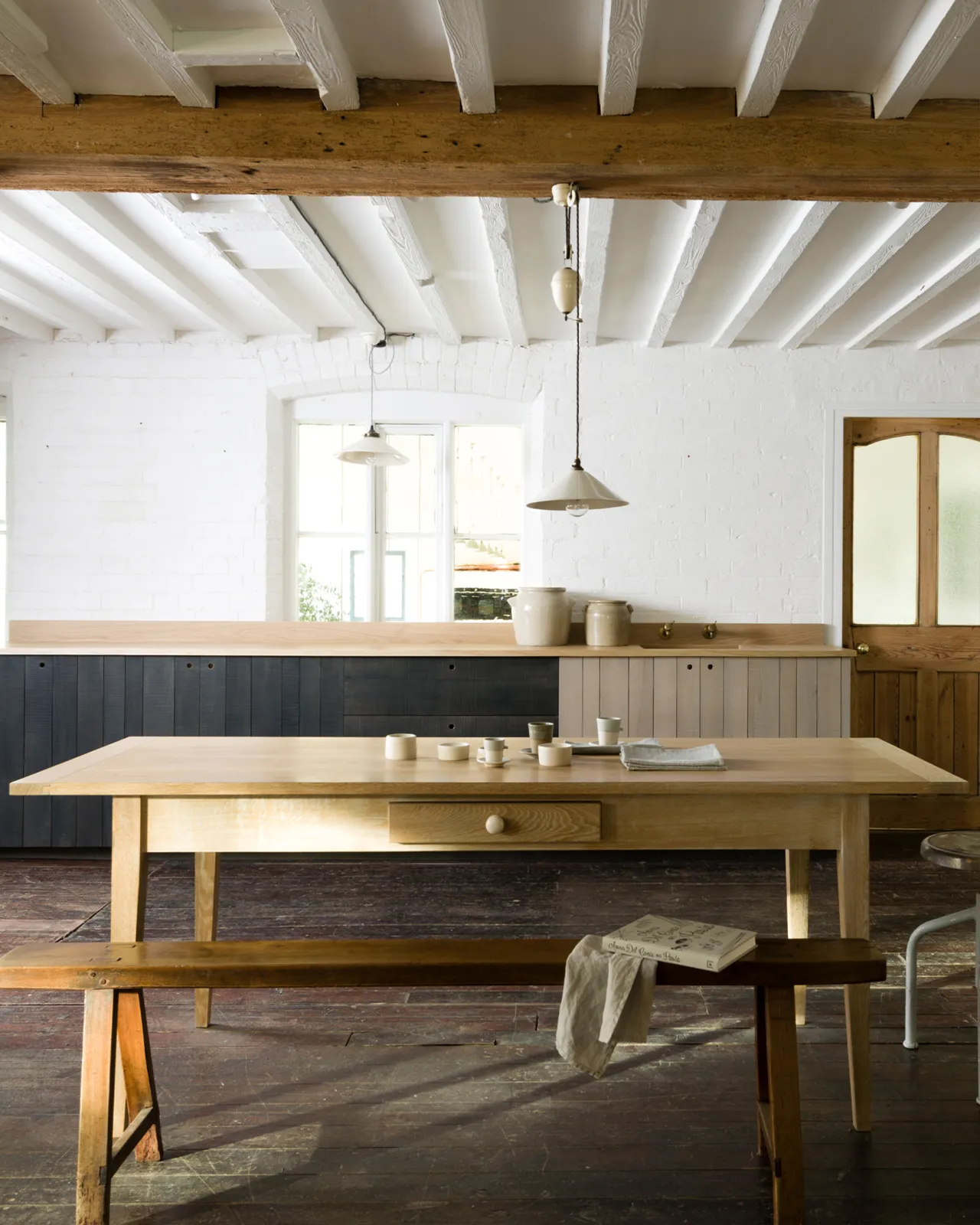 A straight-on view of a Shaker-style oak dining table with breadboard ends, in a cottage room with dark worn floorboards, white brick walls and beamed ceilings.
