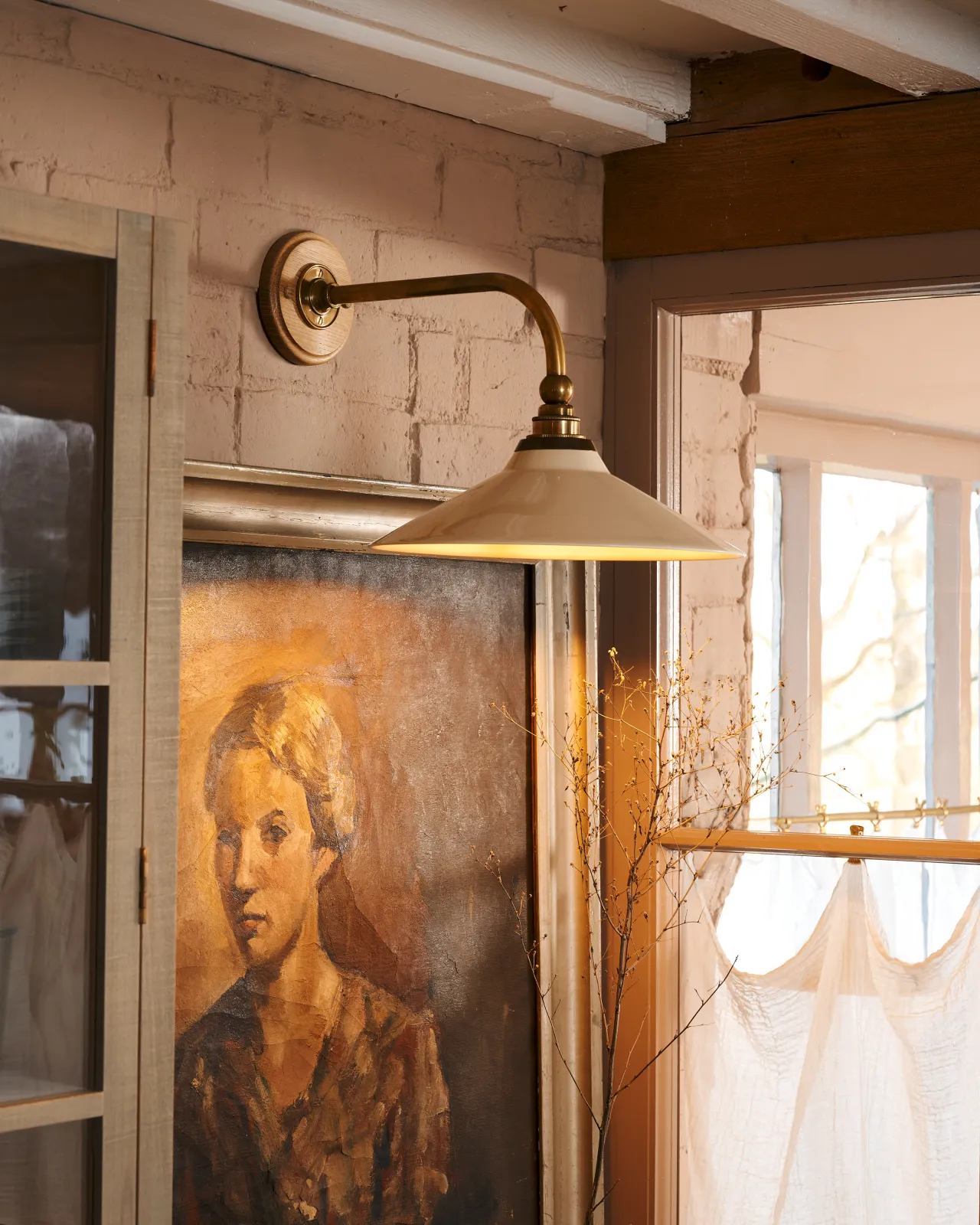 A close-up oblique view of a small Victorian-inspired wall light with a brass arm and a cream glazed ceramic shade fixed to a pink brick wall above an antique portrait painting.