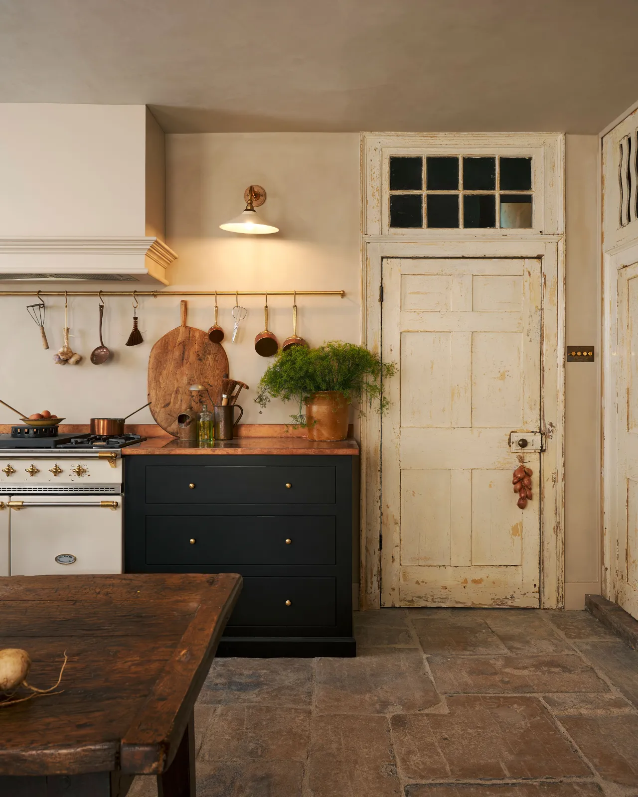 A straight-on view of a rustic kitchen with black painted cabinetry, a white range cooker, stone flooring and cream walls. A small Victorian-inspired wall light with a brass arm and a cream glazed ceramic shade is fixed to the wall beside a closed door.