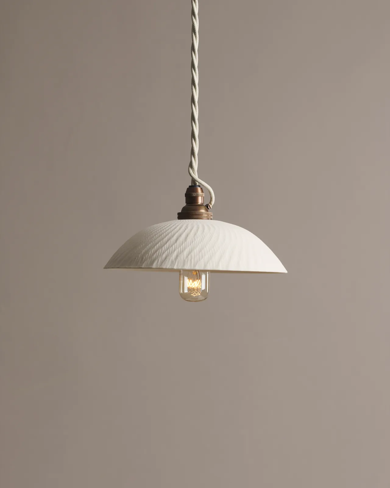 A small chattered skullcap porcelain pendant light with an unglazed finish, an aged brass E27 lampholder and a white twisted fabric flex cord.