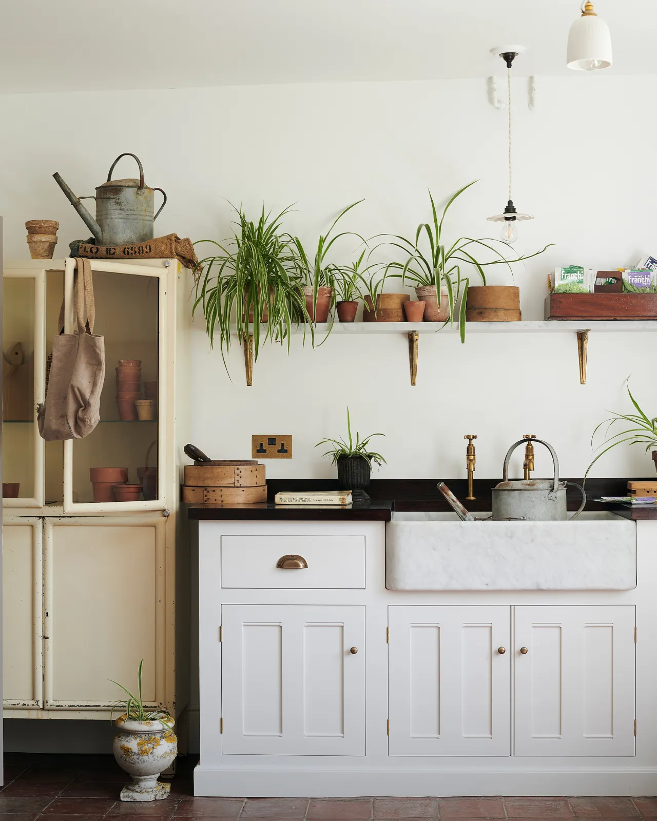 A straight-on view of a classic 13 amp twin plug socket in a mottled aged brass finish in a Mediterannean-inspired kitchen with a white painted Georgian-style sink cabinet, dark hardwood worktop, Carrara marble sink and a shelf full of lush spider plants.