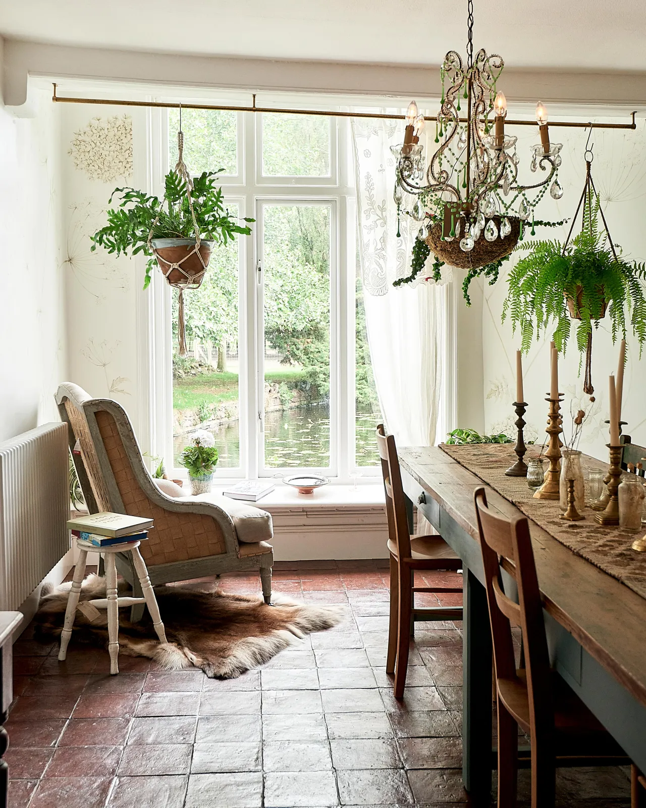 A classically-styled dining room with white painted walls, hanging plants, vintage furniture, square terracotta flooring with an authentic aged finish and a large window with views out into the English countryside.