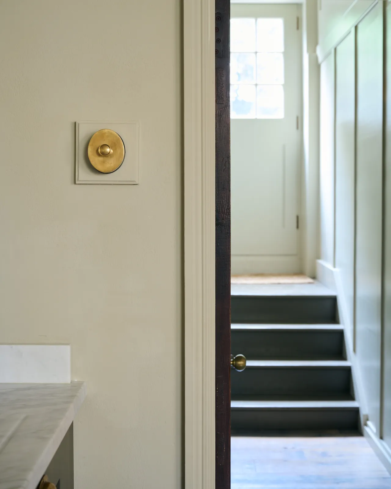 A straight-on view of a small 1 gang oval-shaped dimmer switch in a warm burnished brass finish, mounted on a cream painted square pattress.