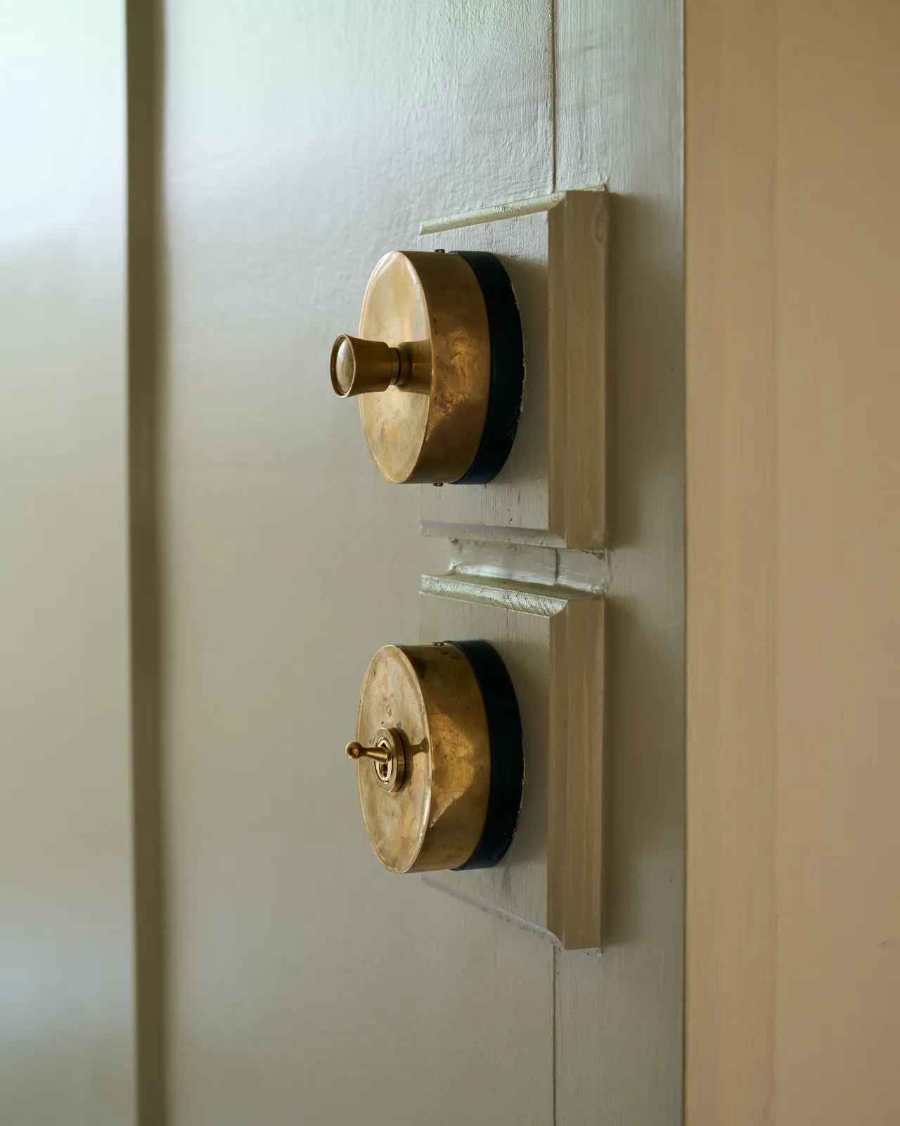 Two small 1 gang oval-shaped switches, one dimmer, one toggle, in a warm burnished brass finish, mounted on glossy cream painted square wooden pattresses.
