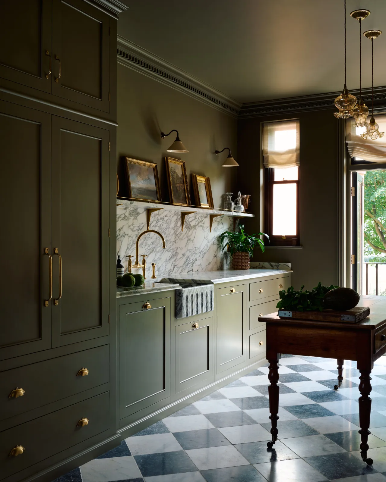 An oblique view of an atmospheric room with dark green painted Georgian-style cabinetry, dark green walls, black and white chequered marble flooring and a row of small wall lights above an Arabescato marble splashback and shelf.