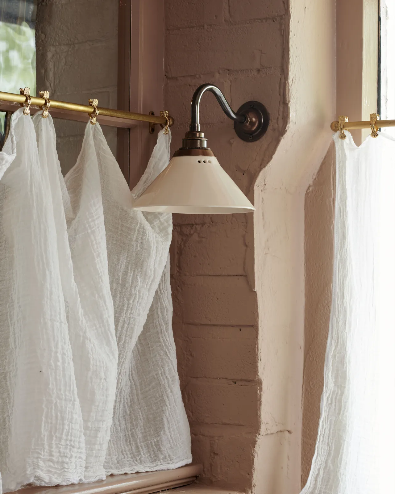 An oblique view of a small wall light with an aged brass swan neck arm and a small cream glazed ceramic shade, fixed to a pink brick wall beside a linen curtain hanging from an aged brass rail.