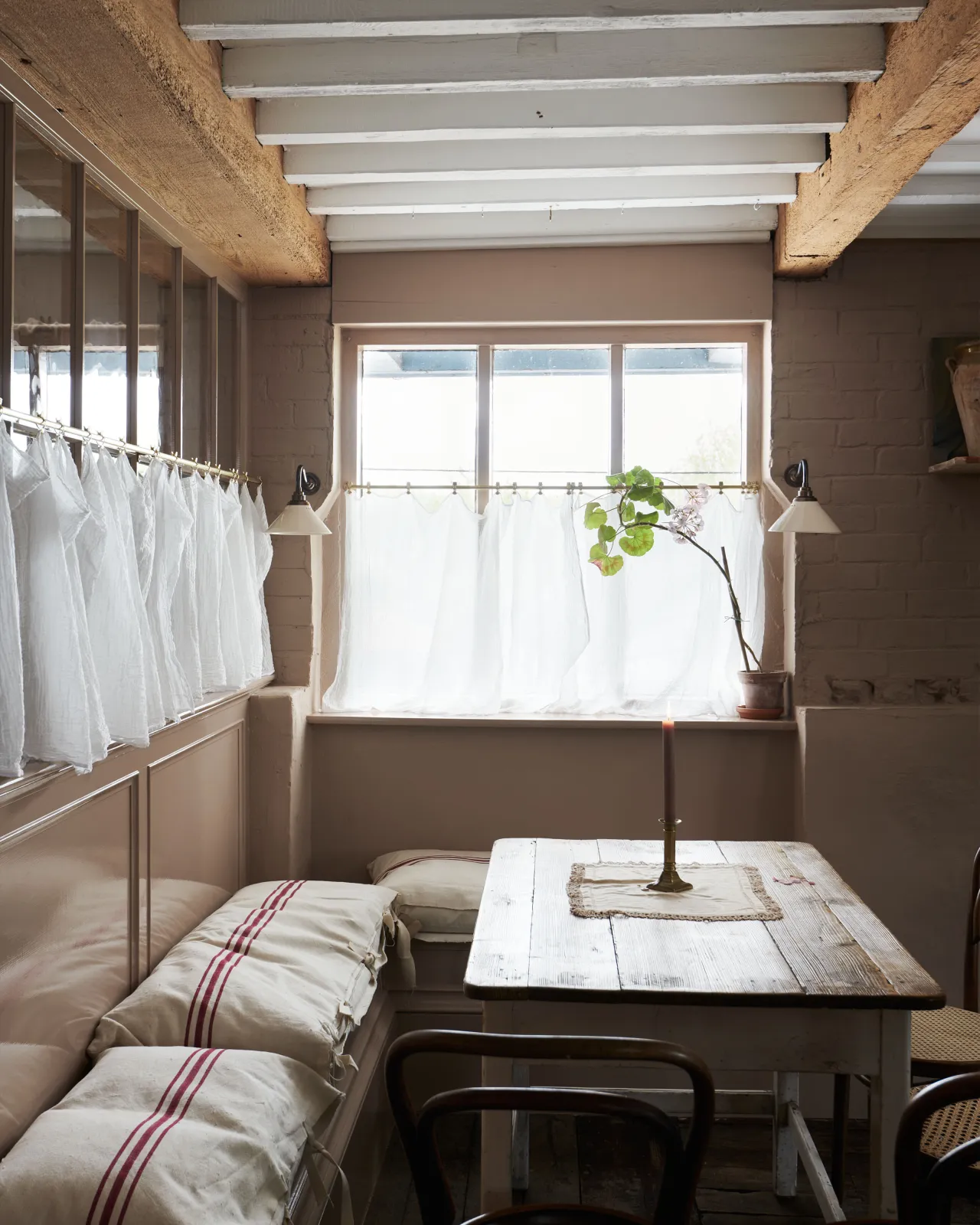 An atmospheric cottage room with pink brick walls, beamed ceiling and small wall lights either side of a window.