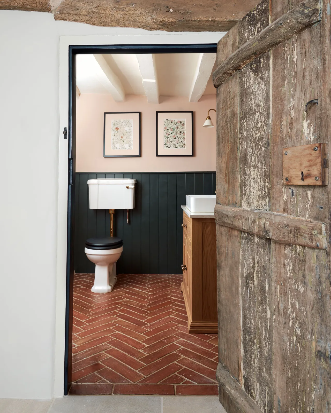Looking through cottage doorway into a small toilet with traditional styling, dark blue painted tongue and groove panelling, soft pink walls, an oak vanity unit and parquet terracotta flooring with an authentic aged finish.