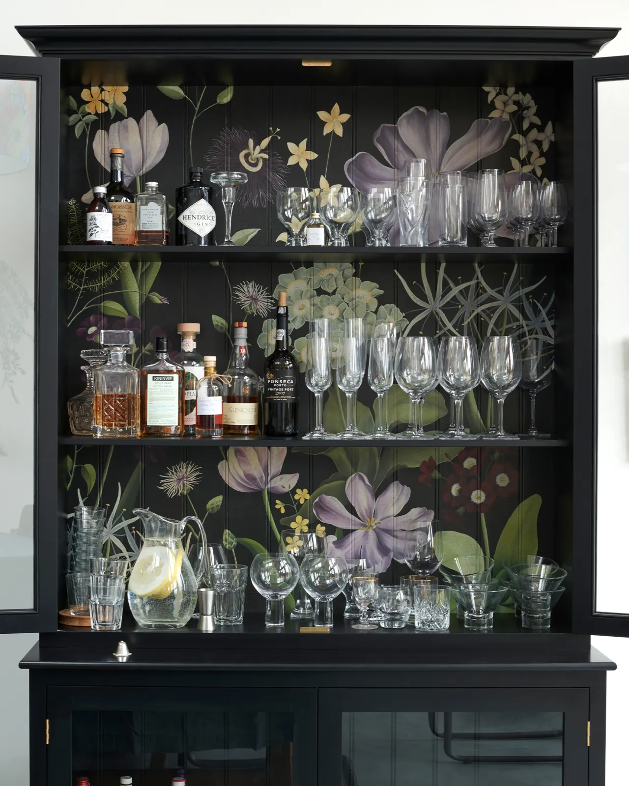 The interior of a black display cabinet with an elegant and bustling painted floral design on a tongue and groove back panel. The shelves are full of clear glasses and bottles of spirits.