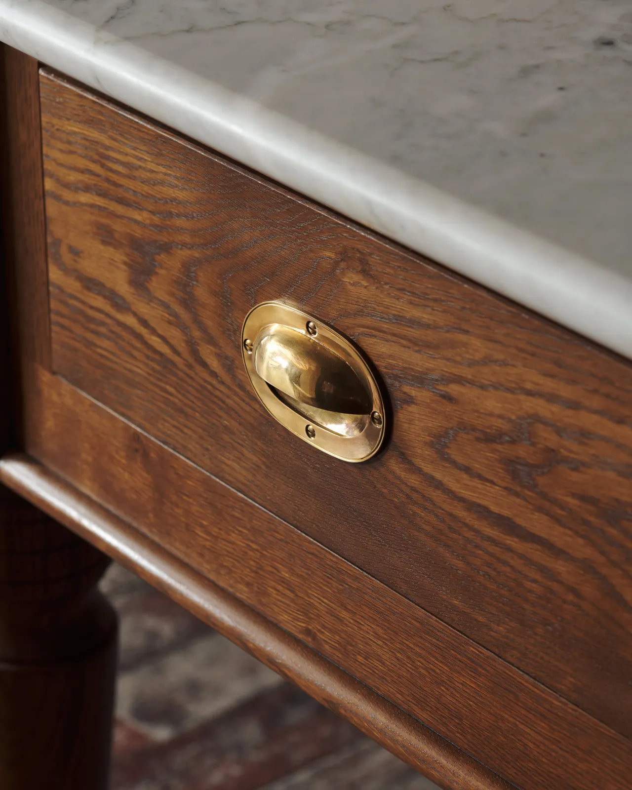 An oblique view of a Victorian-inspired oval-shaped cup handle with an authentic and warm burnished brass finish and a recessed backplate, fitted to a dark stained oak drawer front.