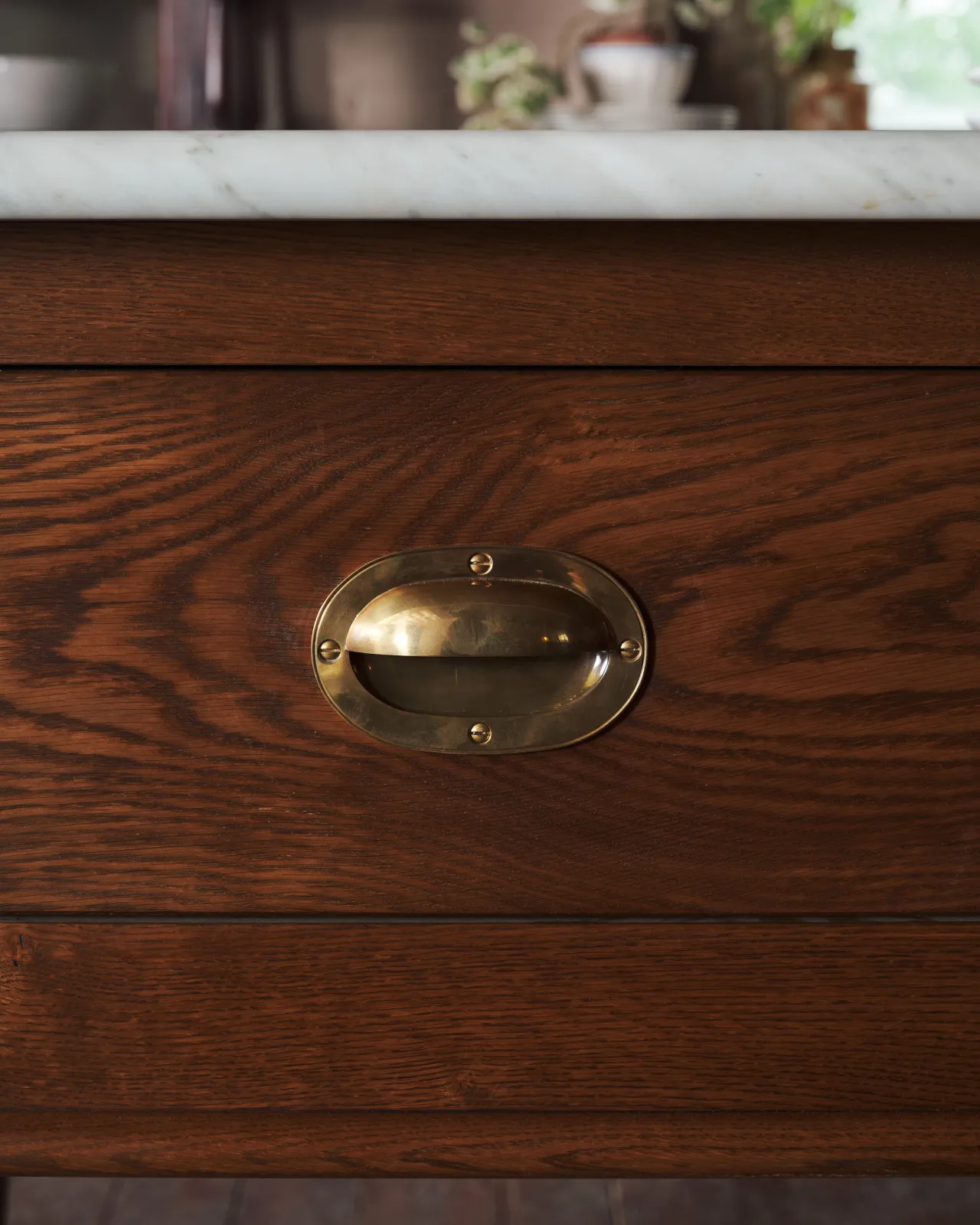 A straight-on view of a Victorian-inspired oval-shaped cup handle with an authentic and warm burnished brass finish and a recessed backplate, fitted to a dark stained oak drawer front.