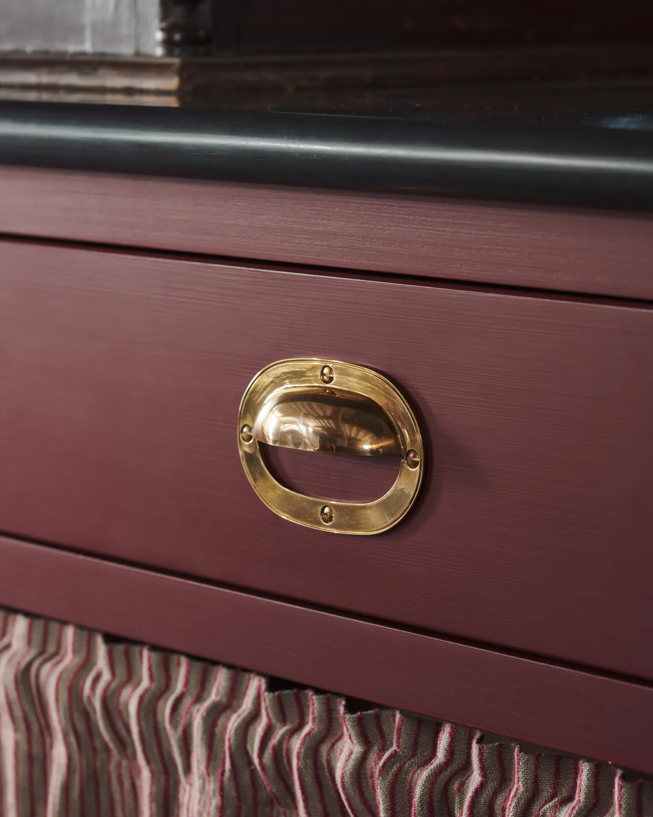 A close-up oblique view of a Victorian-inspired oval-shaped cup handle with an authentic and warm burnished brass finish with no backplate, fitted to a wine red painted drawer front.