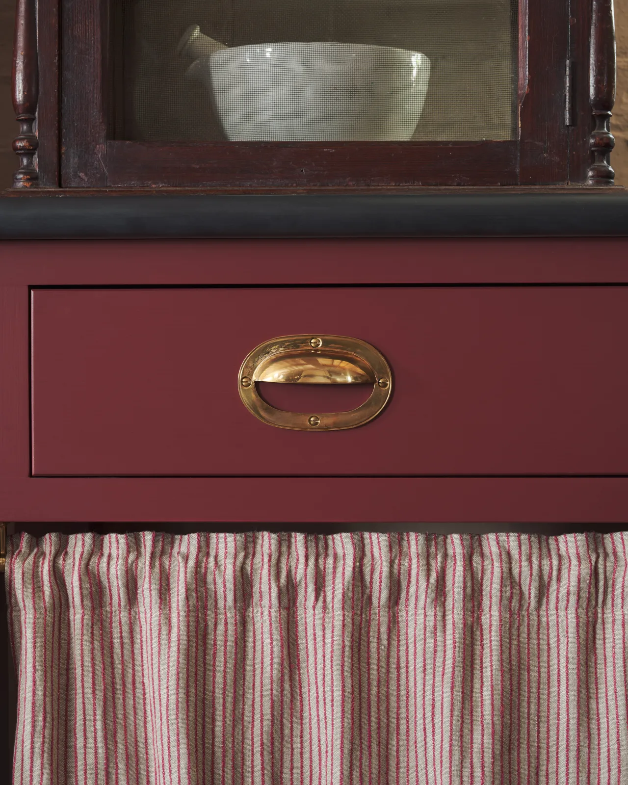 A close-up straight-on view of a Victorian-inspired oval-shaped cup handle with an authentic and warm burnished brass finish with no backplate, fitted to a wine red painted drawer front.