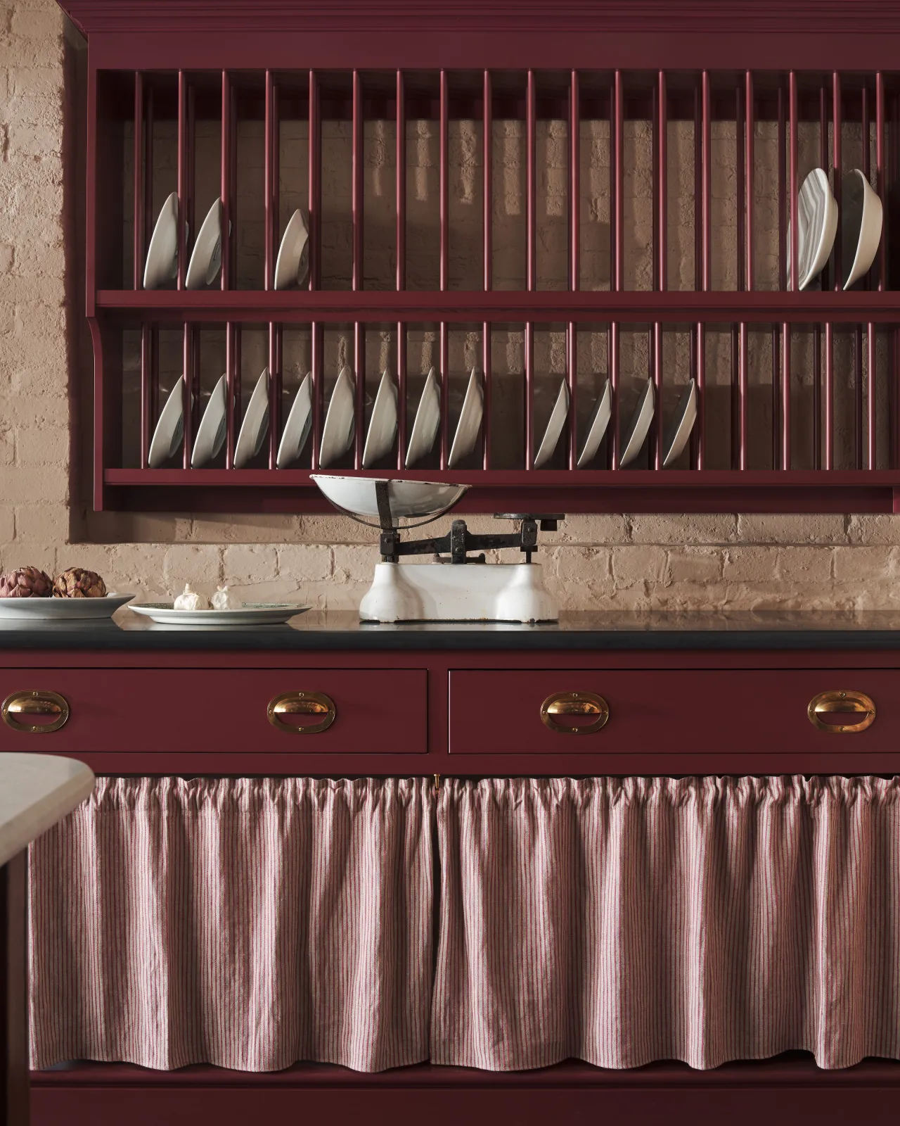 A straight-on view of a Victorian-inspired kitchen with a traditional wall mounted plate rack hanging over a drawer cupboard with open storage behind a striped curtain. The drawers are fitted with oval-shaped cup handles in an authentic and warm burnished brass finish.