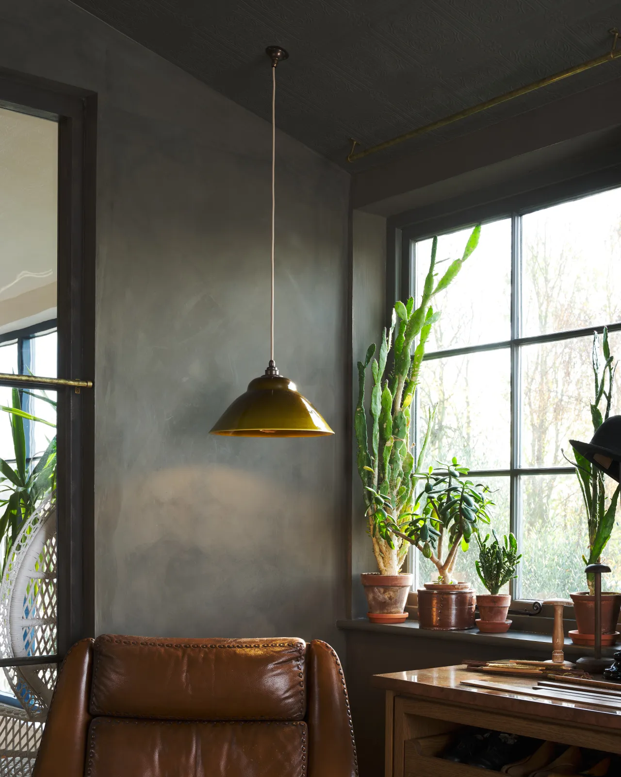 The corner of a dark room with a mid-century-style sepia glazed ceramic pendant light hanging by a window over a brown leather armchair.
