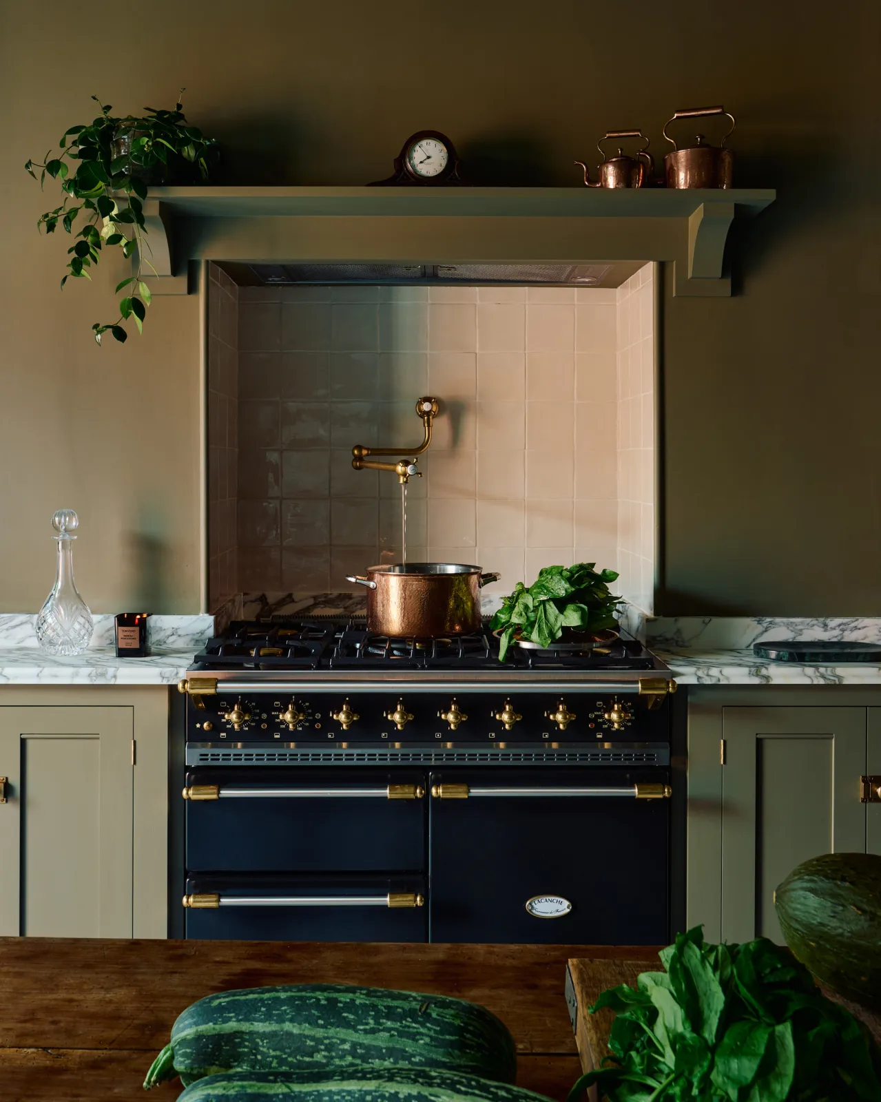A traditional-style aged brass pot filler tap fitted in a small alcove above an impressive black range cooker. The room is painted in a moody dark green colour with an elegant overmantle shelf above the alcove.