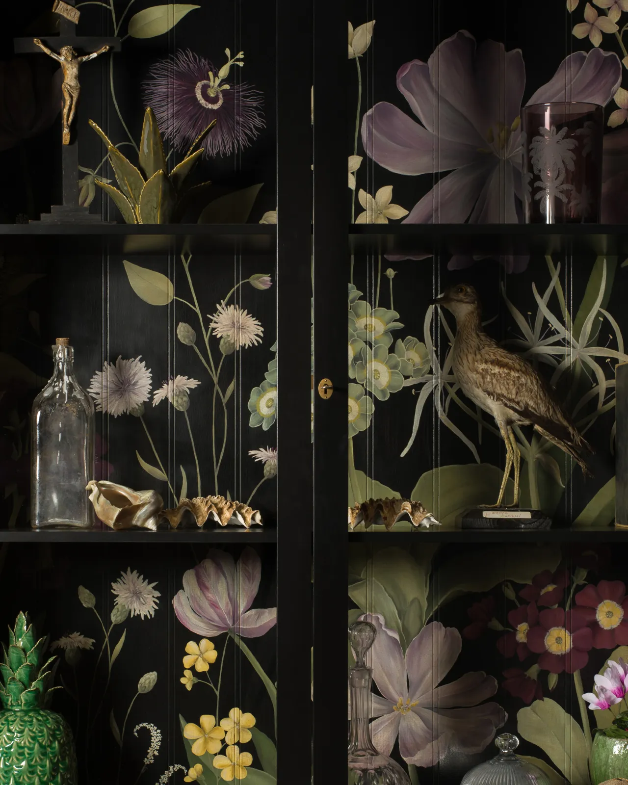 A black display cabinet with glazed doors and an elegant and bustling painted floral design on a tongue and groove back panel. The shelves are full of vintage oddities and taxidermy.