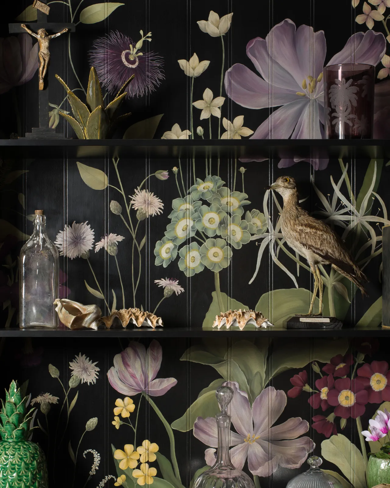 The interior of a black display cabinet with an elegant and bustling painted floral design on a tongue and groove back panel. The shelves are full of vintage oddities and taxidermy.