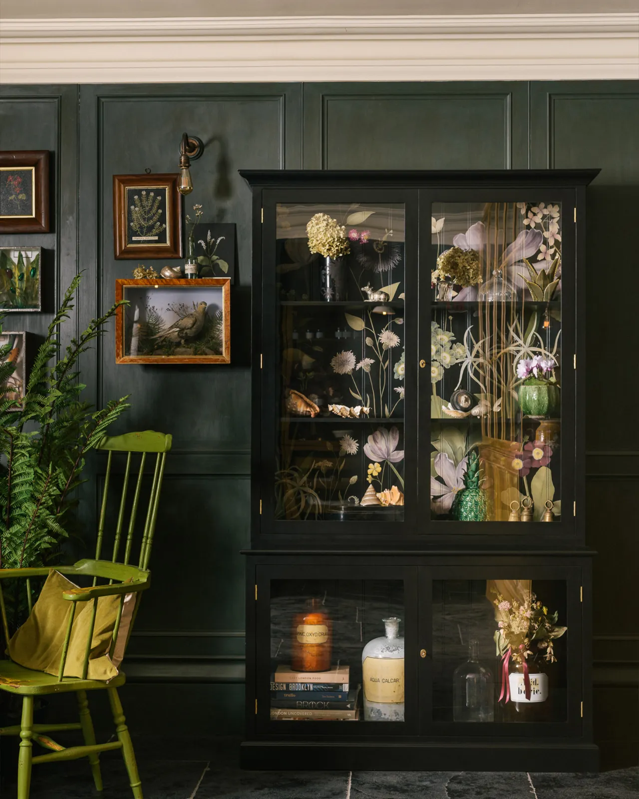 A tall museum-style display cabinet with glazed doors, painted black inside and out and with an elegant and bustling painted floral design on the tongue and groove back panel.