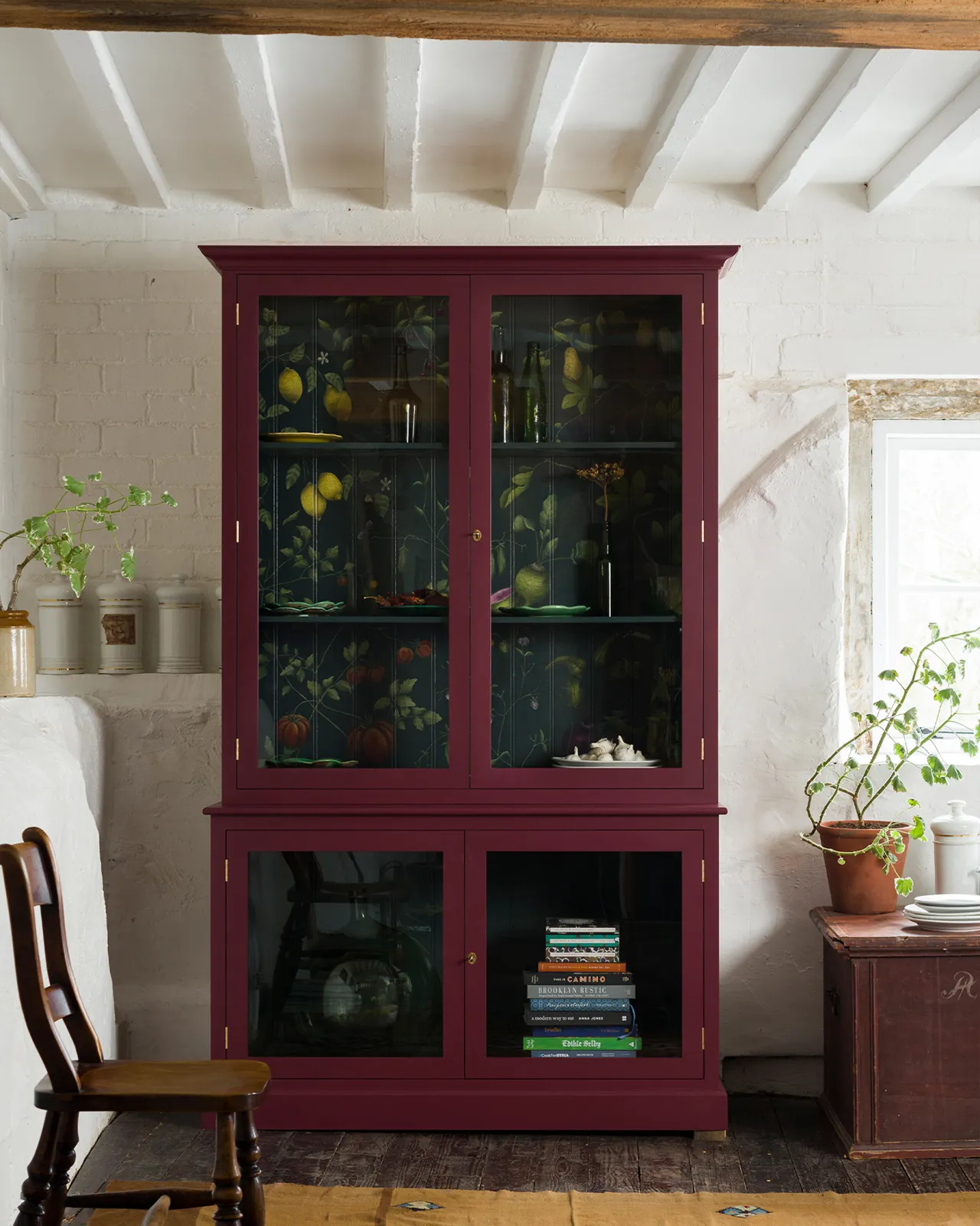 A tall museum-style display cabinet with glazed doors, painted a berry red outside with a dark green interior and a traditional painted botanical design on the tongue and groove back panel.