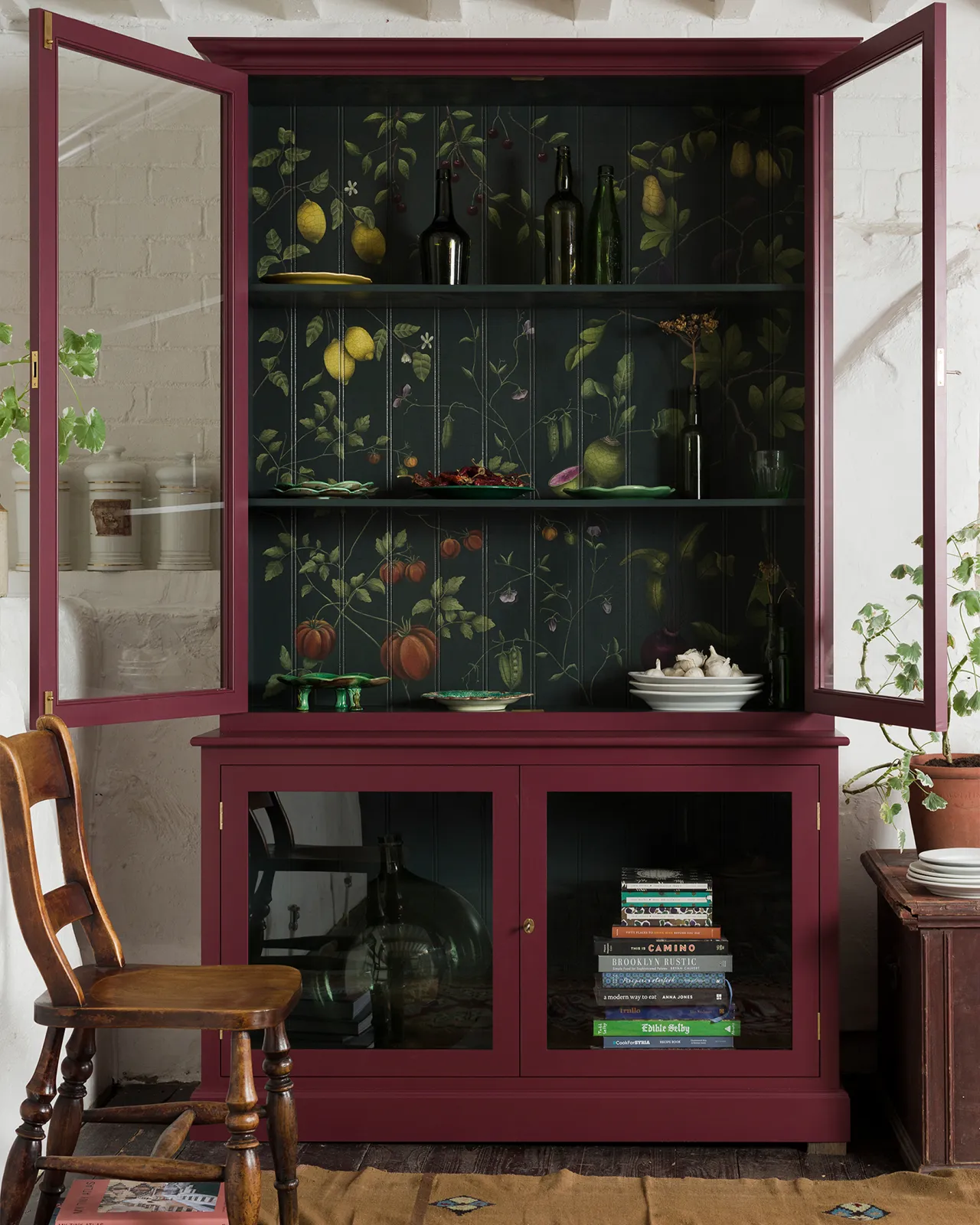 A tall museum-style display cabinet with open glazed doors, painted a berry red outside with a dark green interior and a traditional painted botanical design on the tongue and groove back panel.
