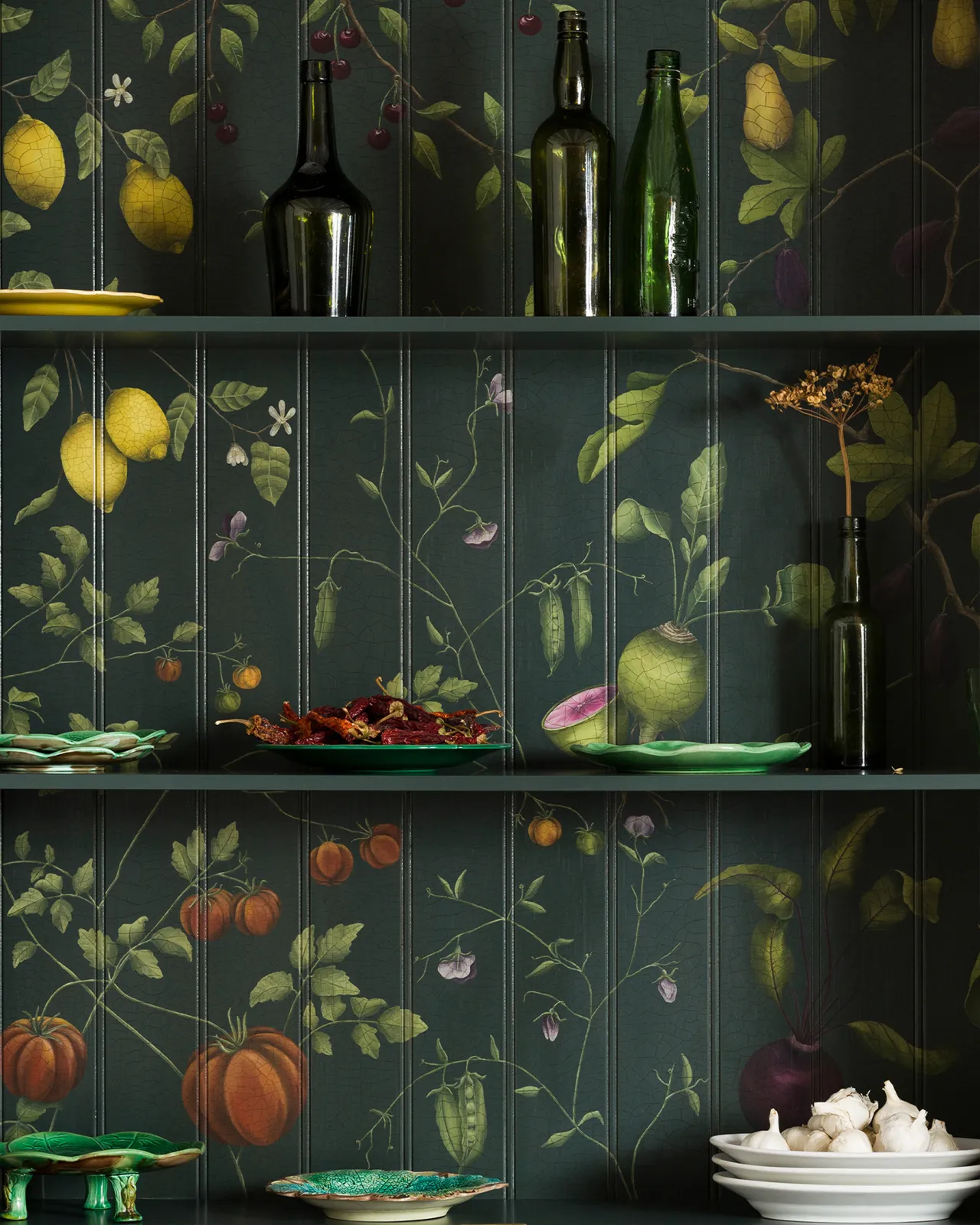 The interior of a display cabinet with a dark green interior and a traditional painted botanical design on the tongue and groove back panel. The shelves are dotted with vintage crockery and green glass bottles.