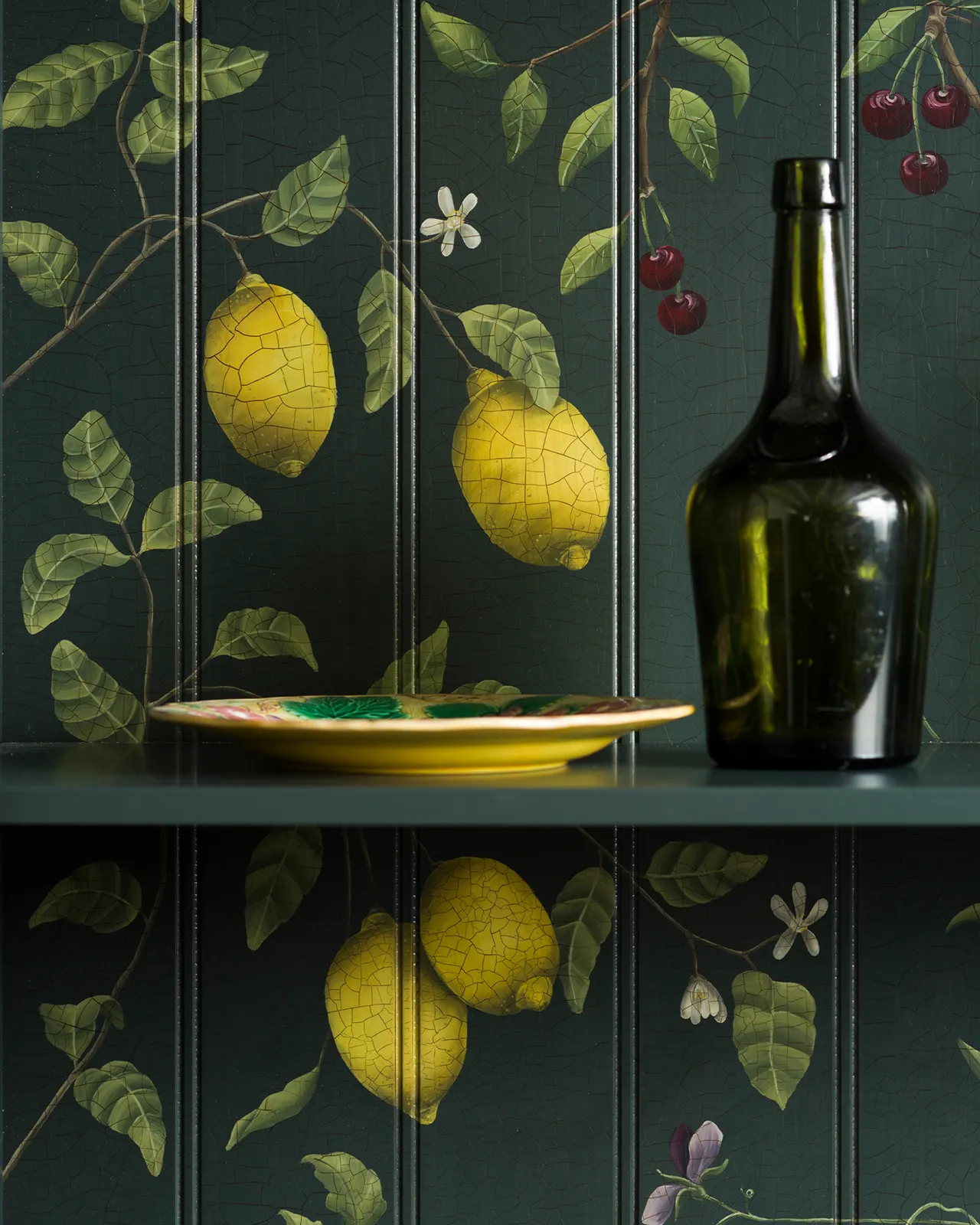 A close-up view of painted lemons and cherries on a dark green tongue and groove back panel. A green glass bottle sits on a shelf in front.