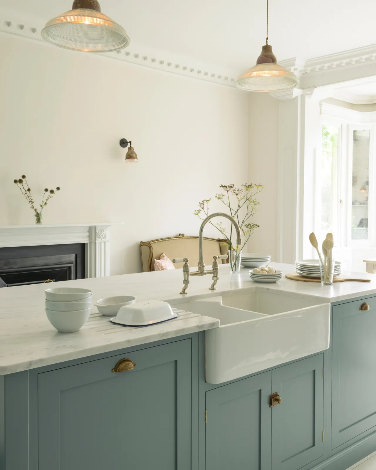 An oblique view of a duck-egg blue painted Shaker-style kitchen sink cabinet with a ceramic Belfast sink, Carrara marble worktop and white walls.