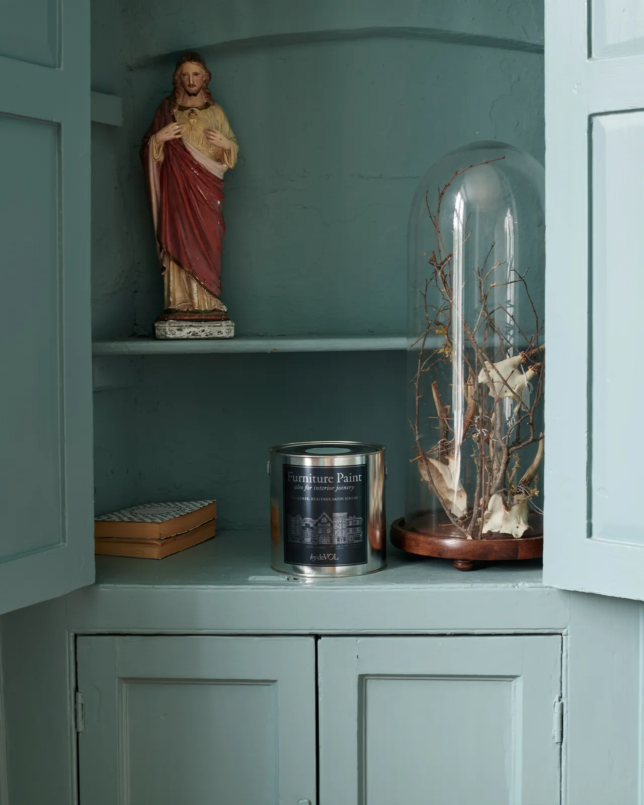 A silver paint tin with a blue label in an open duck-egg blue painted fitted cupboard with a ceramic statue of Jesus and a tall glass bell jar terrarium.