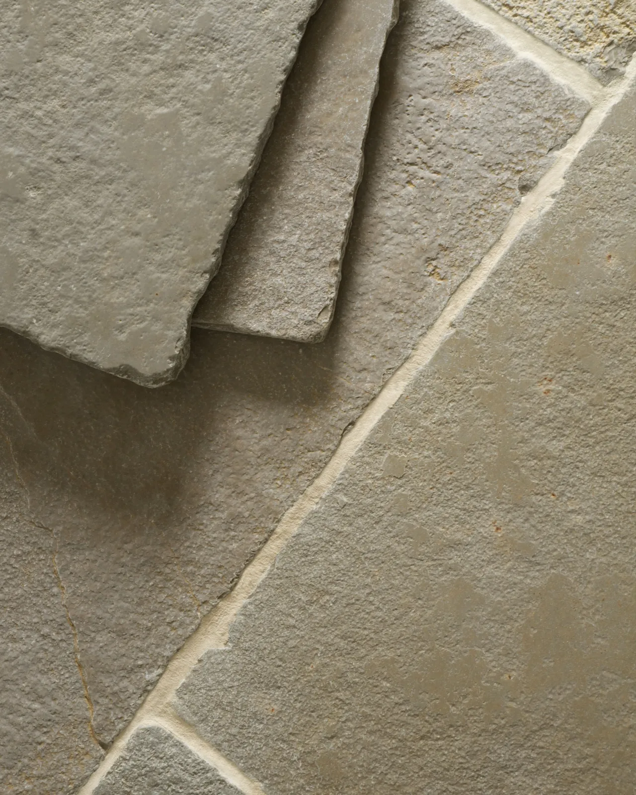 A close-up overhead view of an aged limestone floor with natural earthy tones of green, brown and grey, with two loose tiles stacked in one corner.