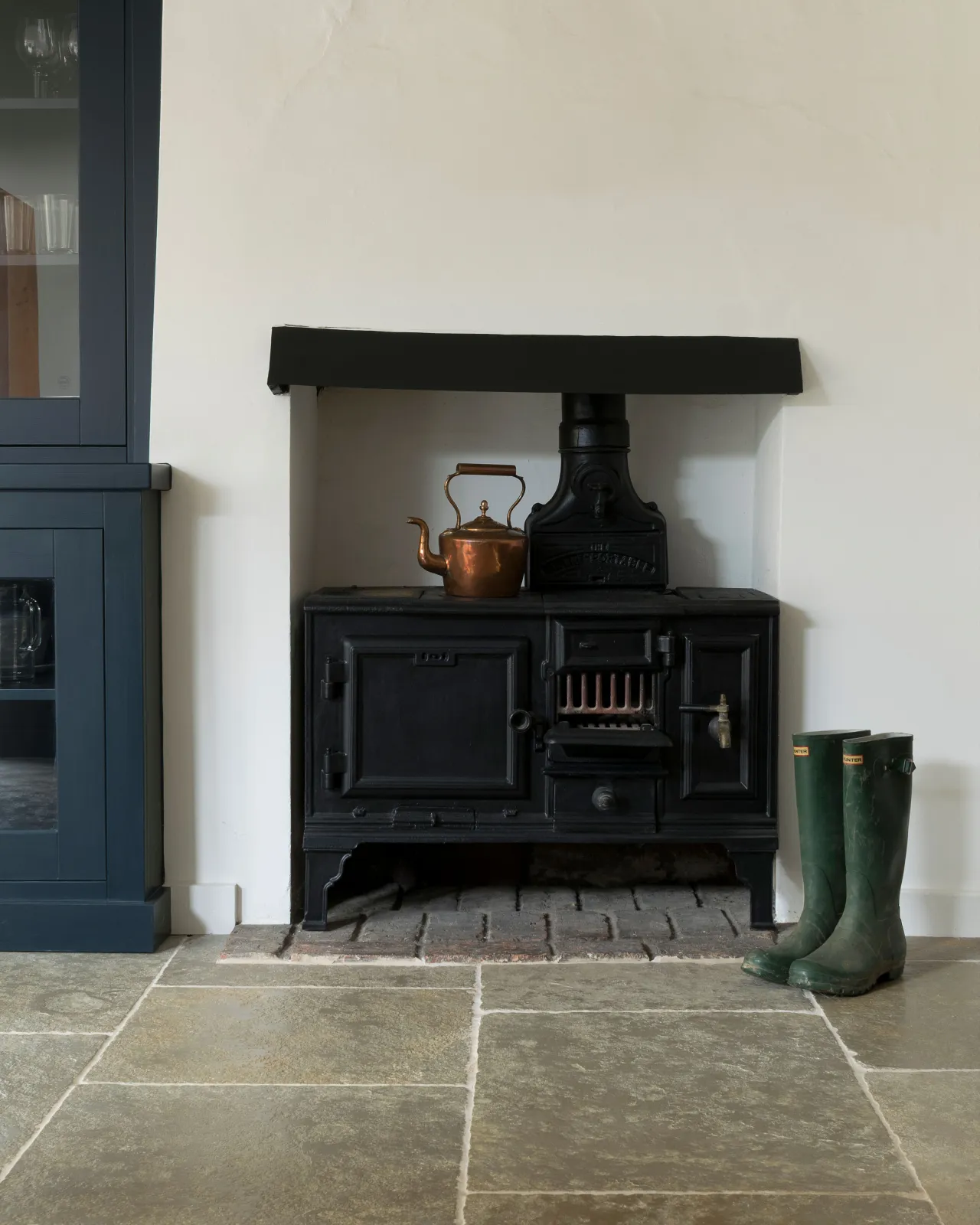 A black antique stove cooker in a small alcove with white walls, green wellington boots and an aged limestone floor with natural earthy tones of green, brown and grey.
