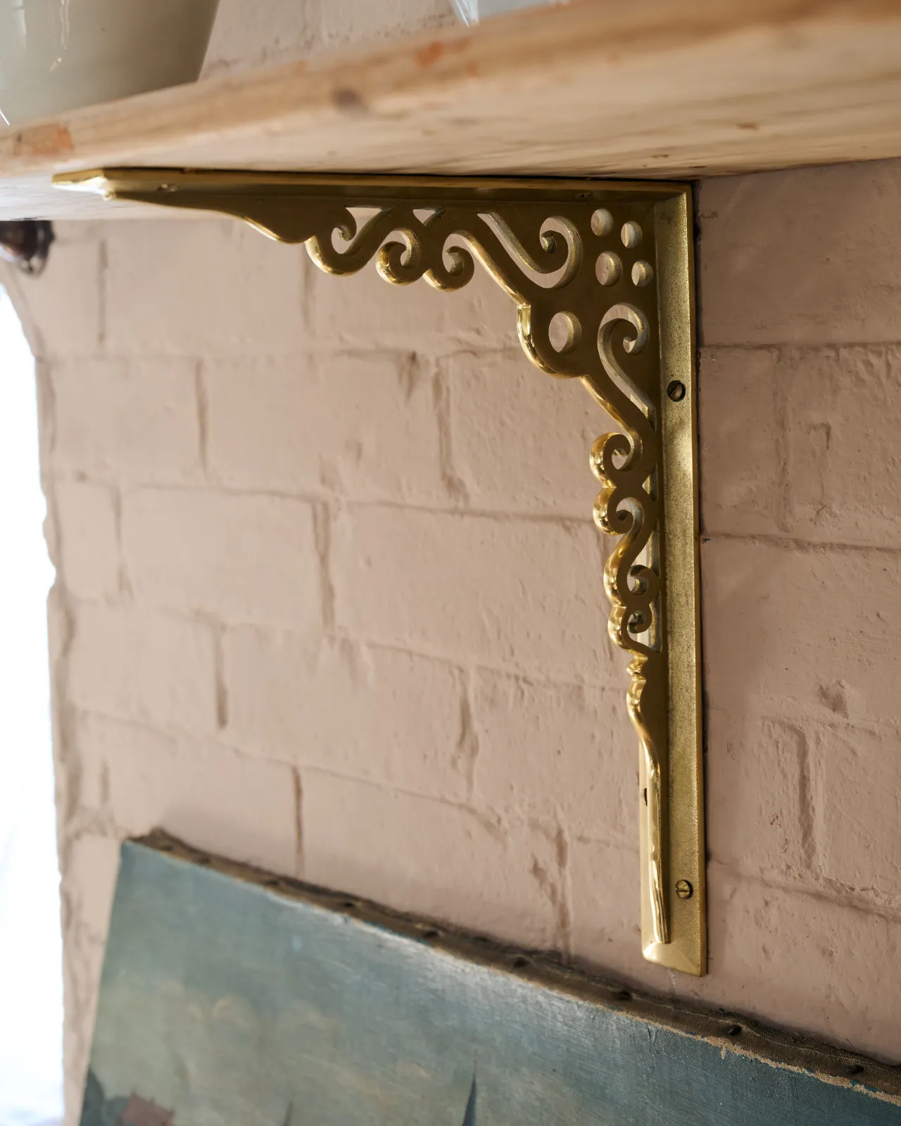 An oblique view of a large Victorian-inspired ornate aged brass shelf bracket holding a deep pine shelf to a pink colour brick wall.