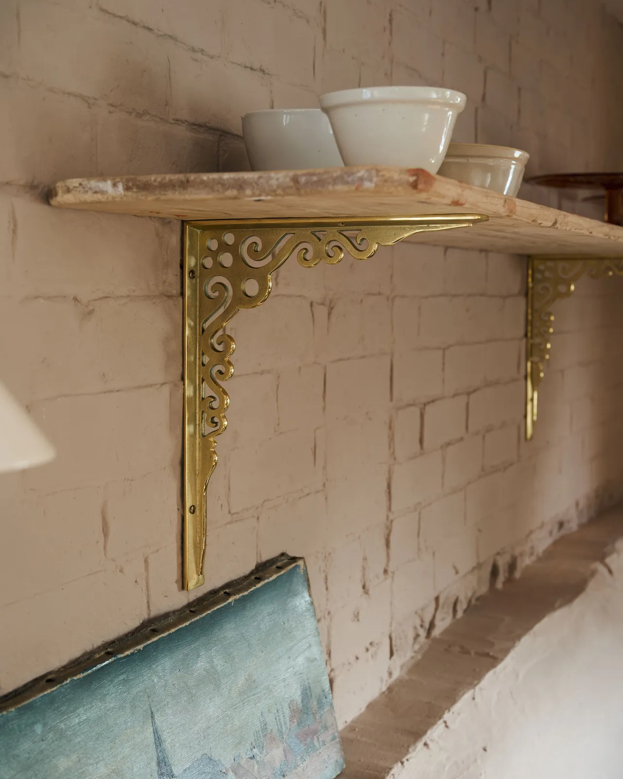An oblique view of a large Victorian-inspired ornate aged brass shelf bracket holding a deep pine shelf to a pink colour brick wall.