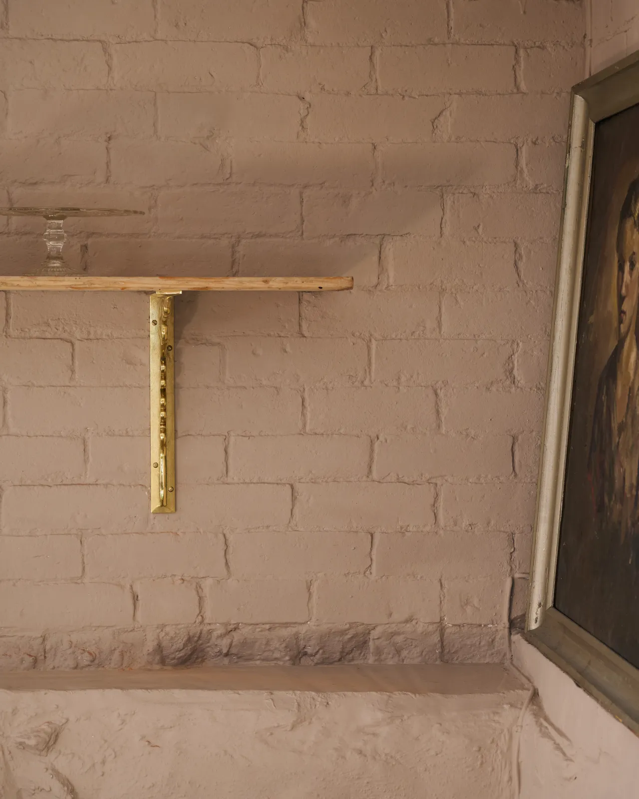 A straight-on view of a large Victorian-inspired ornate aged brass shelf bracket holding a deep pine shelf to a pink colour brick wall.