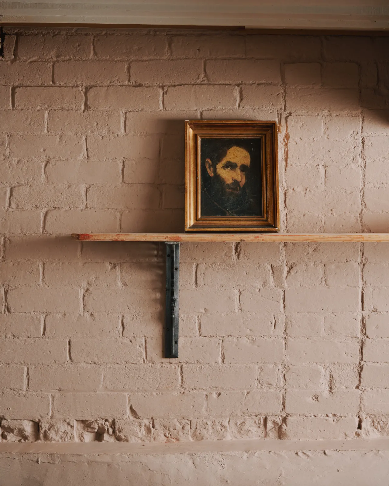 A straight-on view of a large Victorian-inspired ornate cast iron shelf bracket holding a deep pine shelf to a pink colour brick wall.