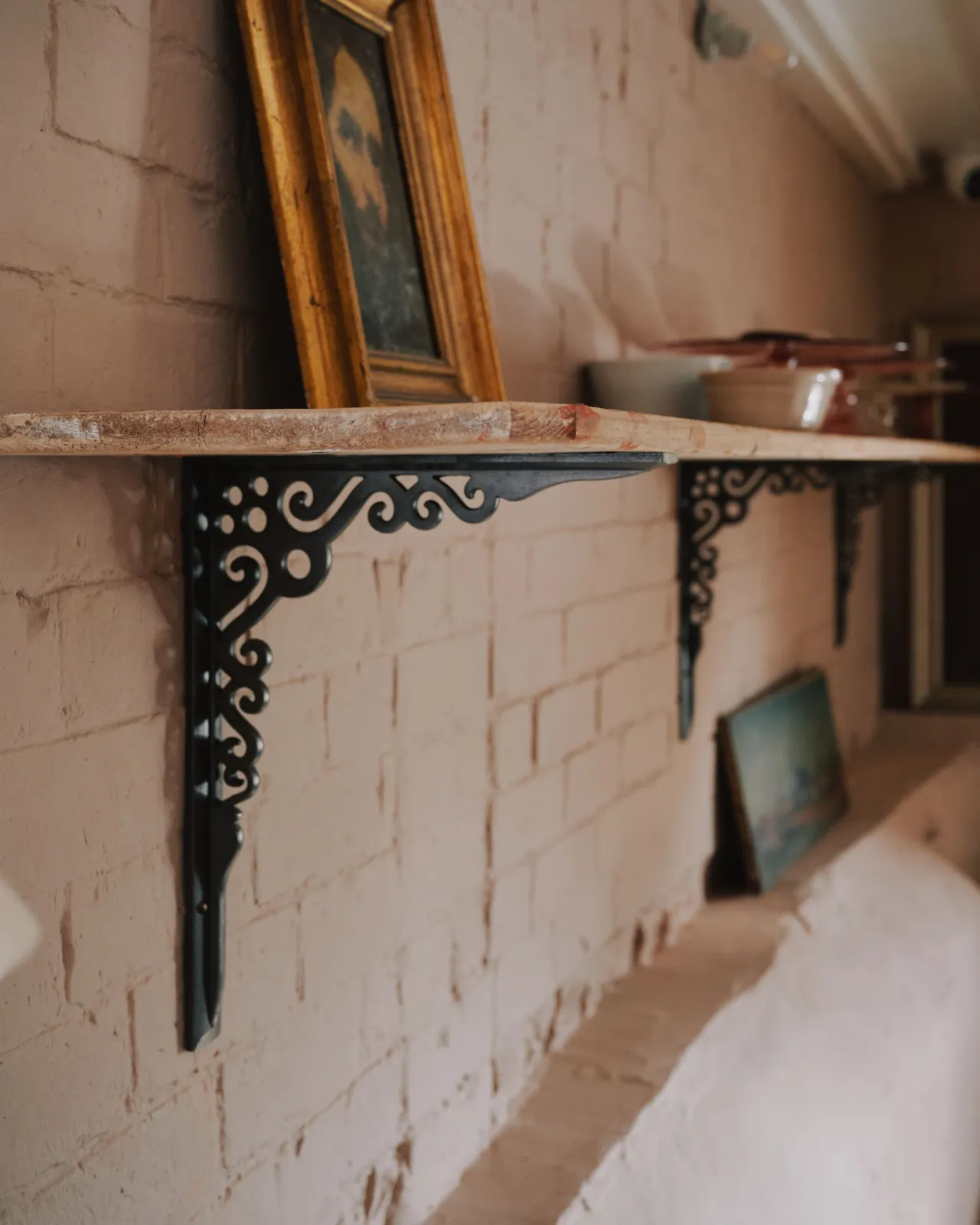 An oblique view of a large Victorian-inspired ornate cast iron shelf bracket holding a deep pine shelf to a pink colour brick wall.