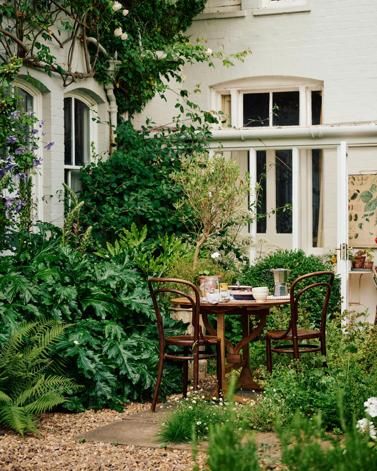 A small round pine dining table and two vintage café-style chairs outdoors in the corner of a lush cottage garden with bushy ferns.