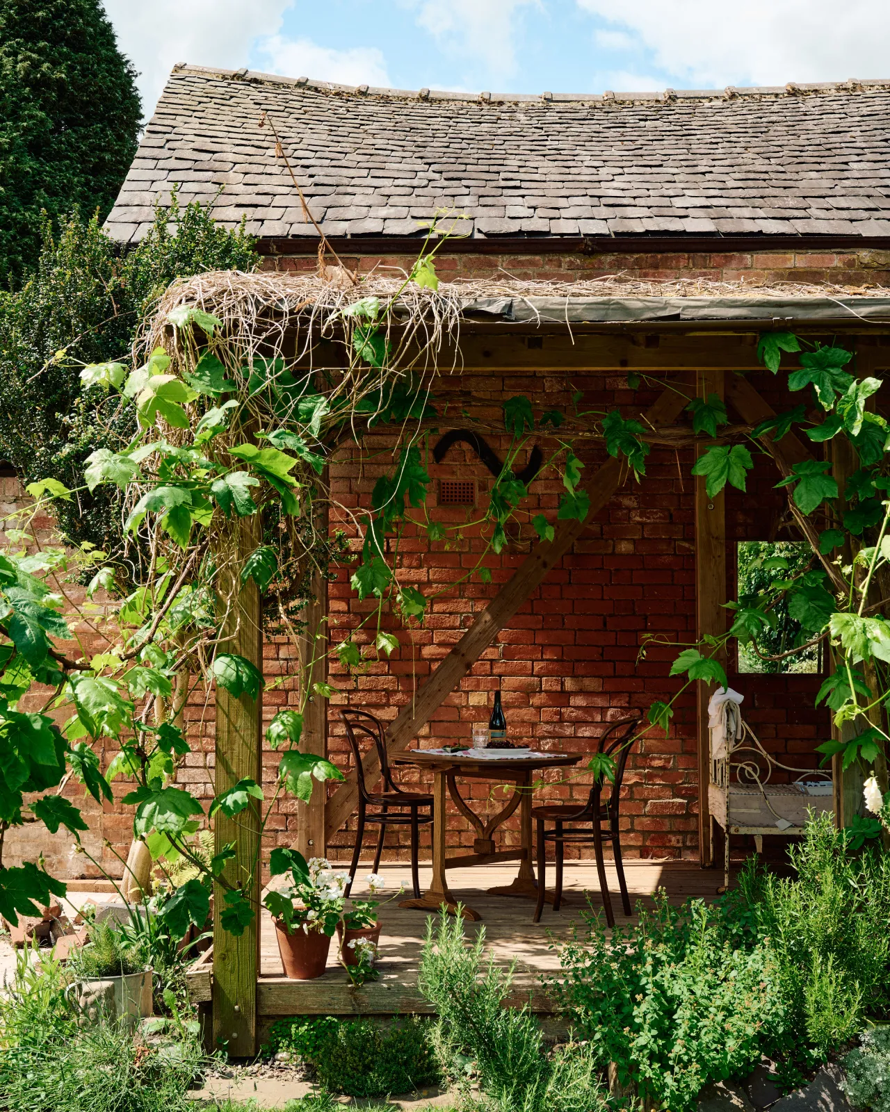 A small round pine dining table and two vintage café-style chairs outdoors on wooden decking, surrounded by lush climbing vines.