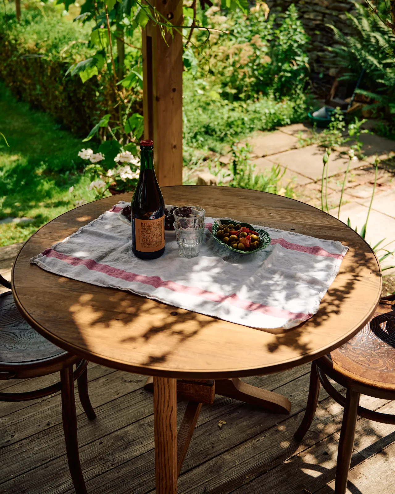 An oblique view of a small round pine dining table with a small vintage linen tablecloth and a spread of red wine, olives and cherries.