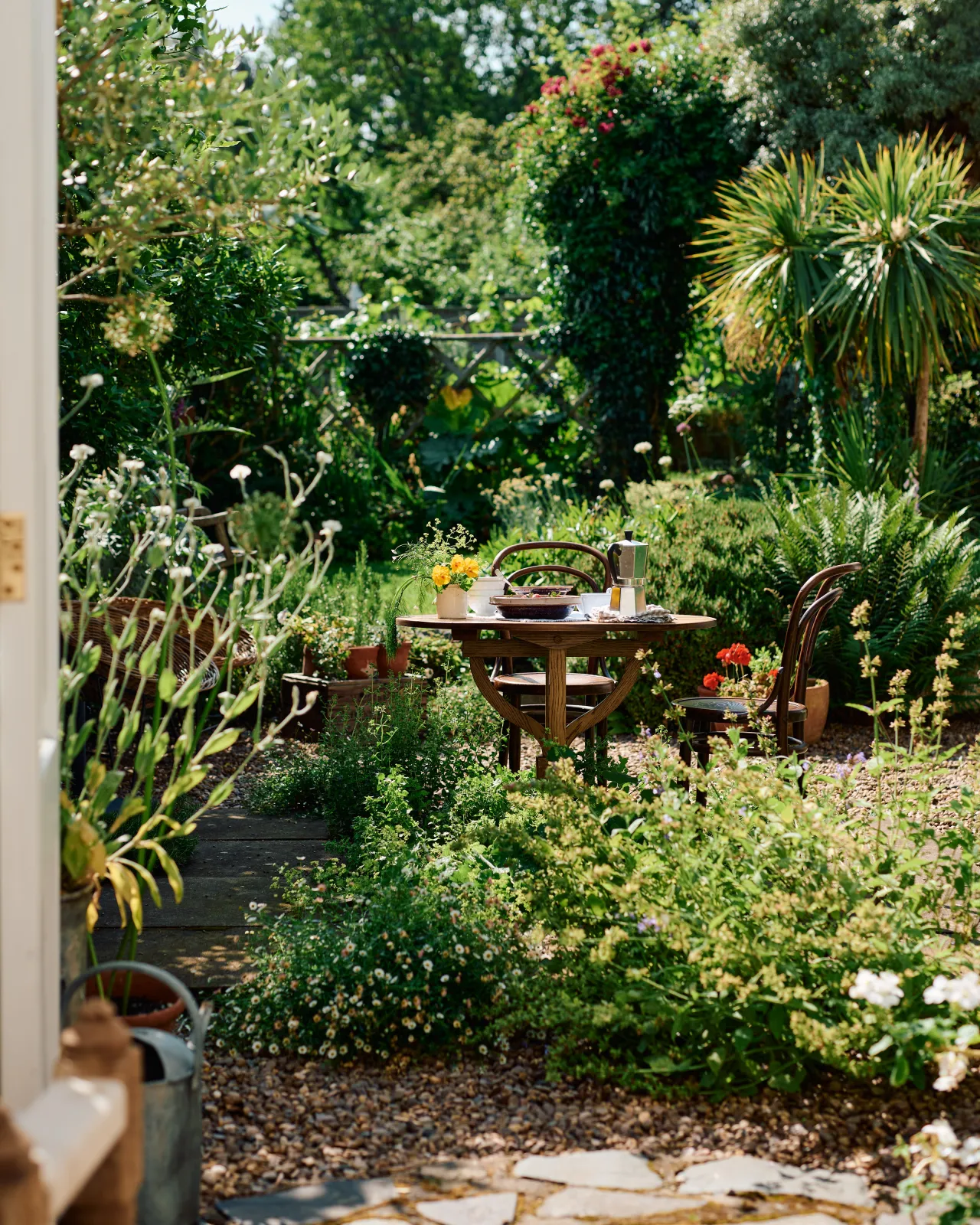 A small round pine dining table and two vintage café-style chairs outdoors in a lush summer garden with wild flowers and ferns.