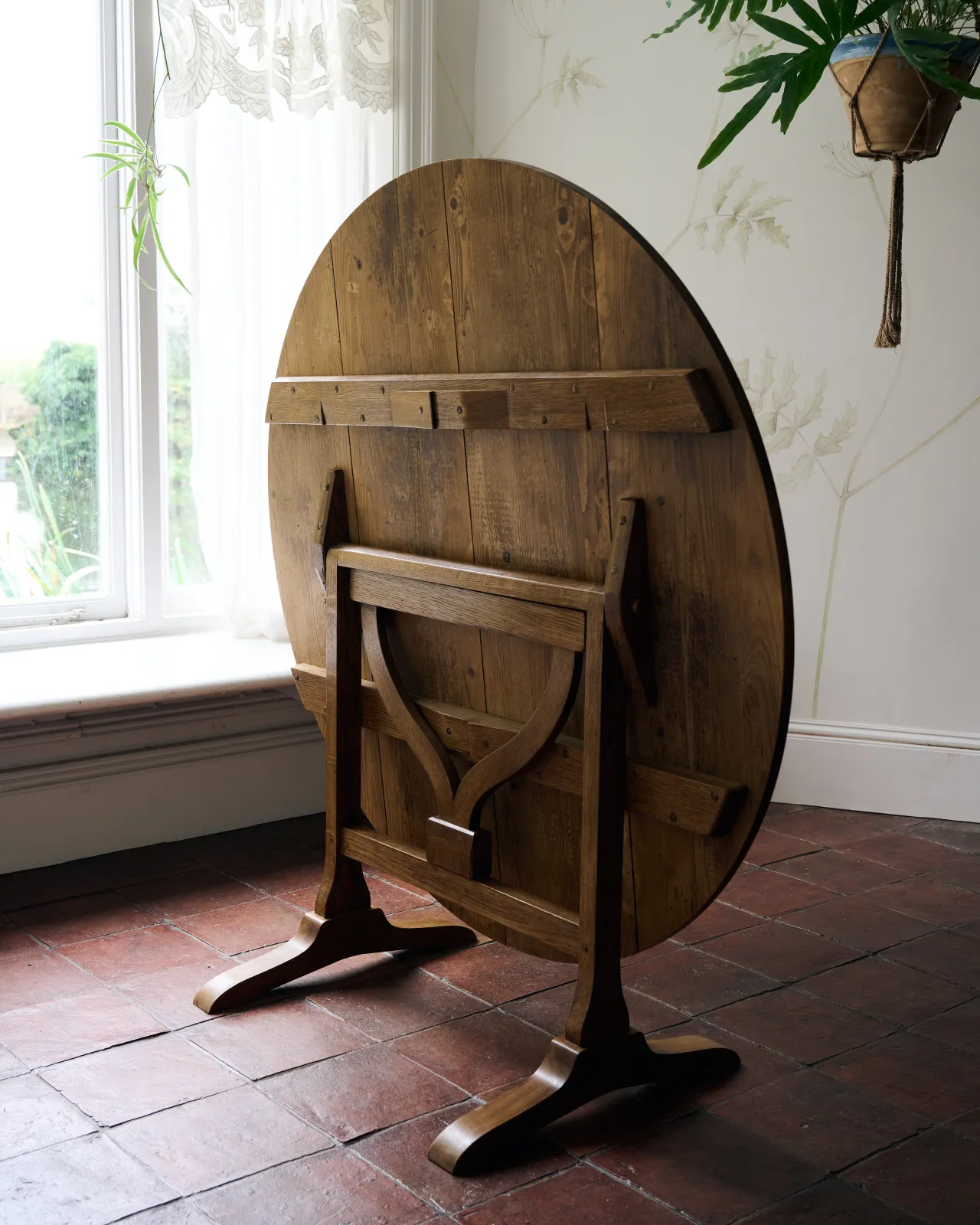 An oblique view of a round pine dining table which has the top folded upright to reveal a simple turning support mechanism built into the frame below.
