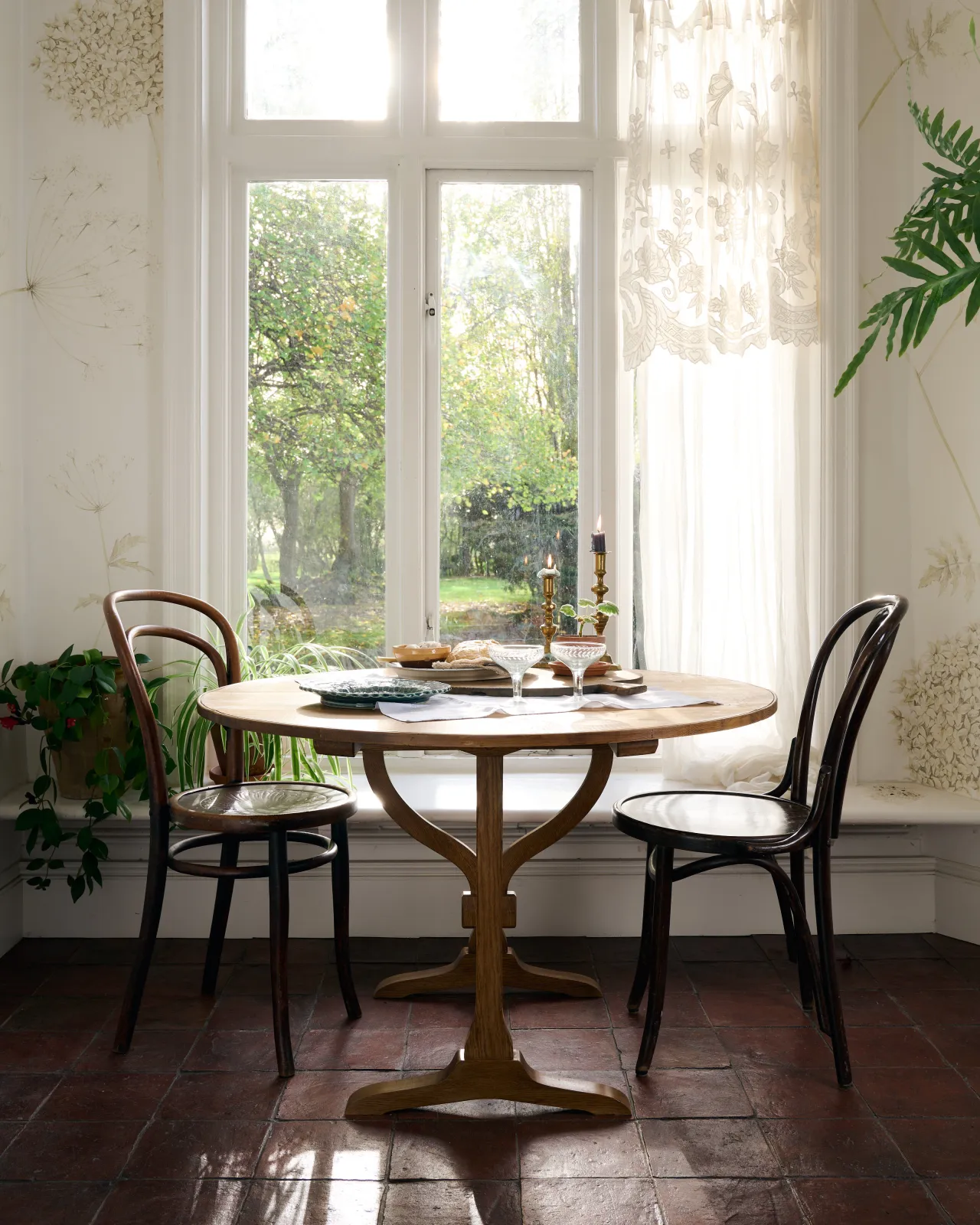 A straight-on view of a round pine dining table with two café-style chairs in front of a tall Victorian cottage window with views over a millpond.