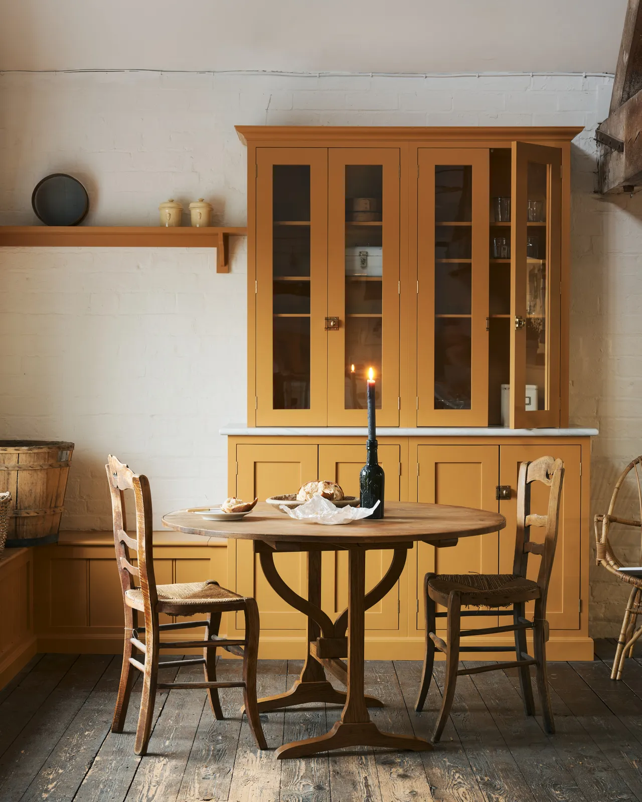 A straight-on view of a round pine dining table with vintage wooden chairs, in a rustic cottage kitchen with yellow painted Shaker-style cabinetry, dark worn floorboards, white brick walls and wooden beams.