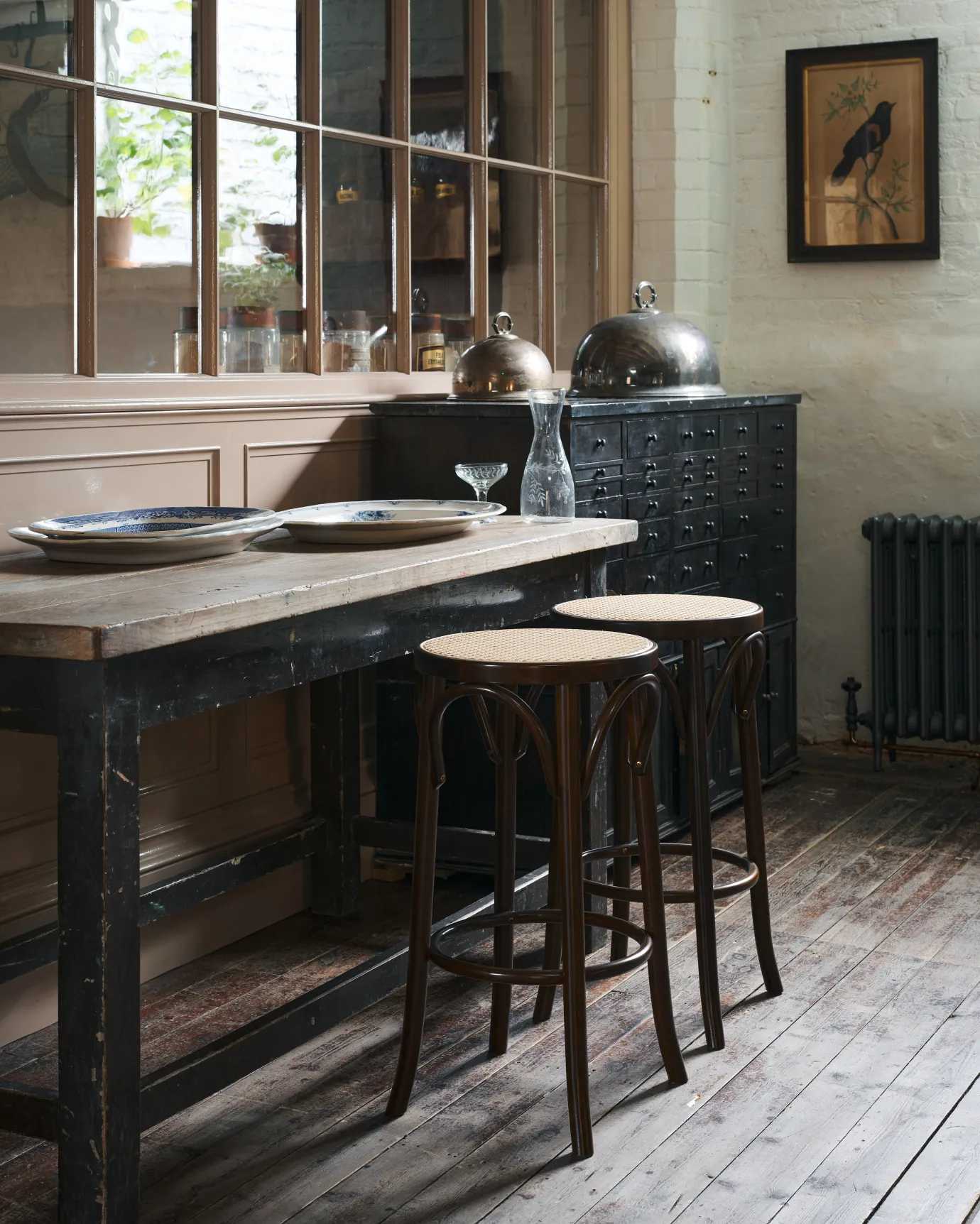 An oblique view of two café-style bar stools with dark stained frames and light woven ratten seats by a vintage table.