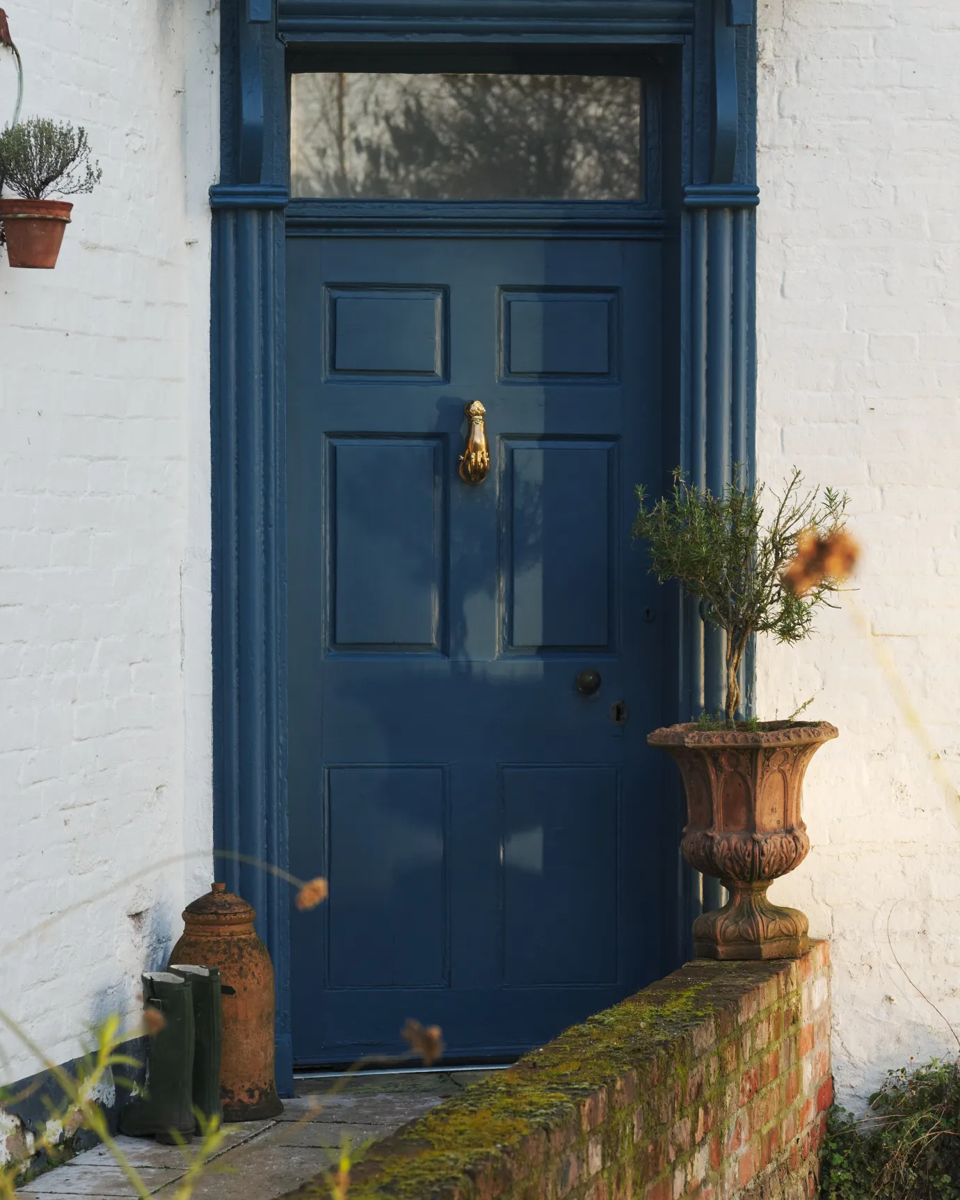 A straight-on view of a cottage with white brick walls and a blue painted front door and a decorative brass door knocker, shaped like a hand holding a pomegranate.