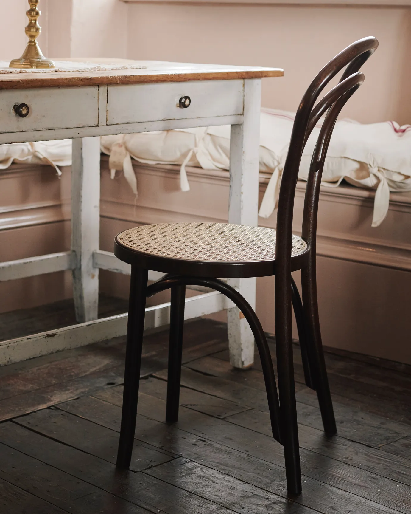 An oblique view of a café-style bentwood chair with a dark stained frame and light woven rattan seat mesh in front of a vintage pine table.