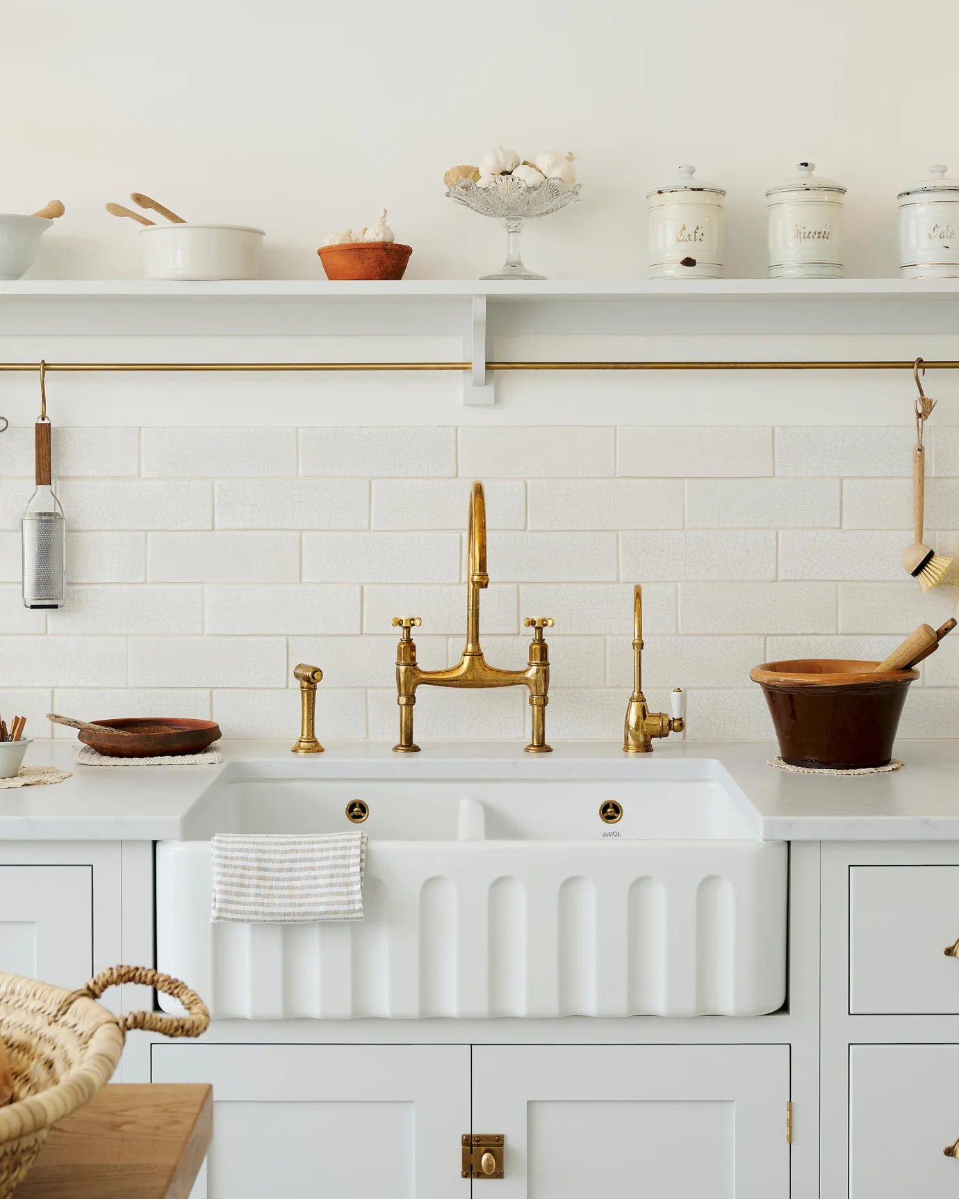 A straight-on view of a large fluted double bowl ceramic Belfast sink in a white painted simple Shaker-style kitchen with an aged brass mixer tap and white metro wall tiles.