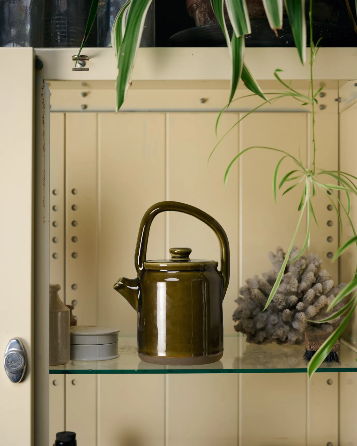 A small mid-century-style ceramic teapot with a glossy amber glaze in a cream coloured medicine cabinet with glass shelving.