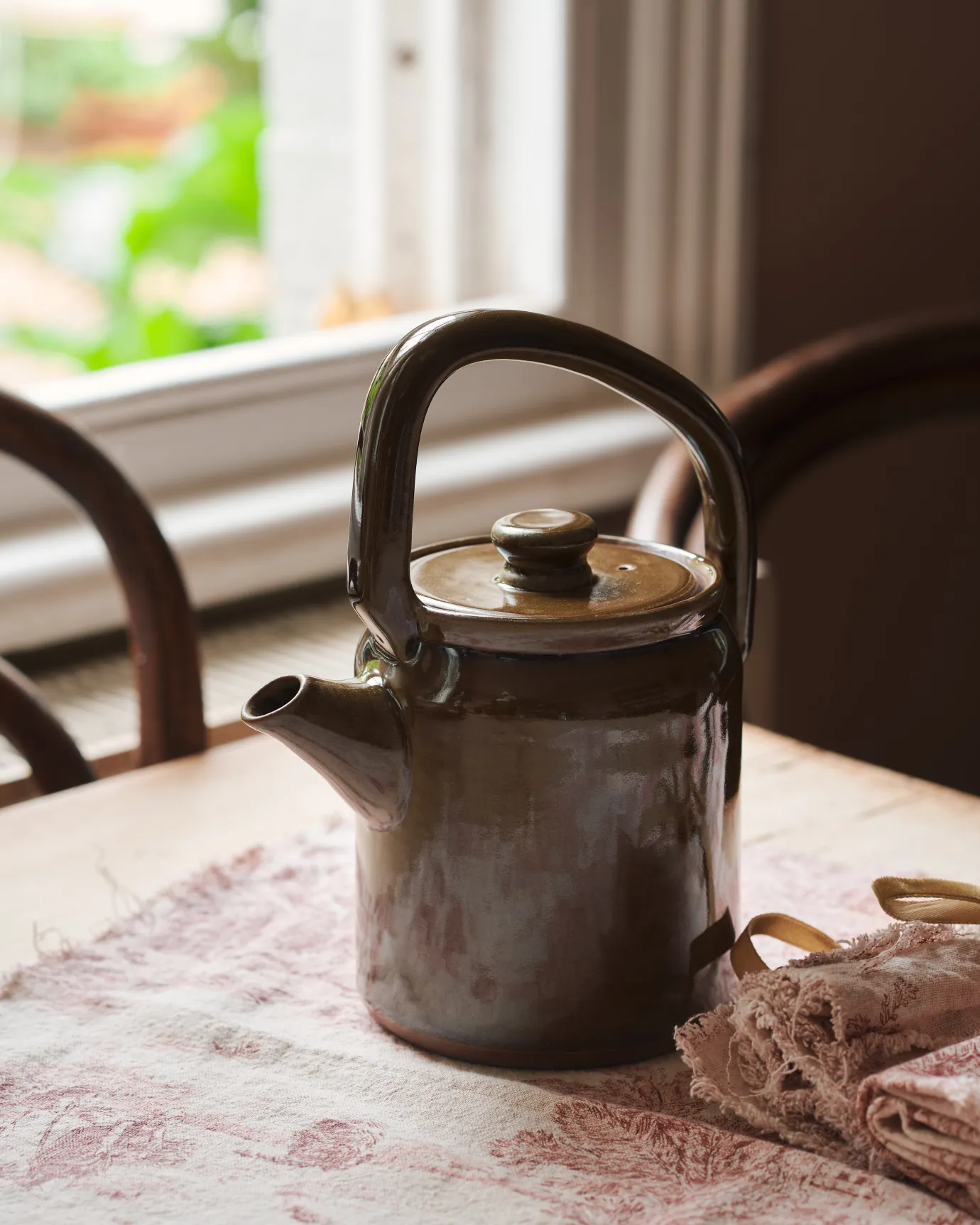 A small mid-century-style ceramic teapot with a glossy amber glaze on a table with a vintage tea towel.