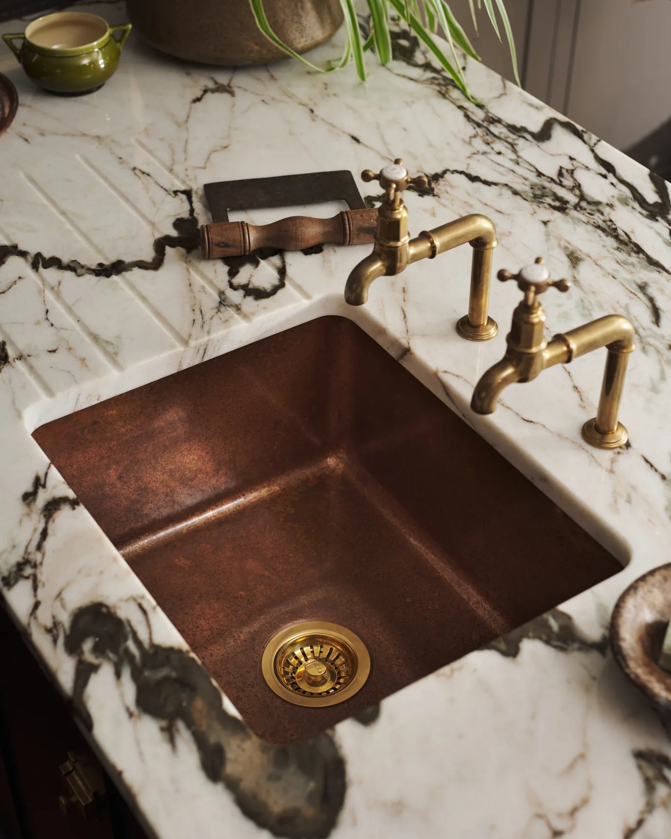 Angled top view of the copper sink with brass taps and a marble countertop, featuring a small green bowl and leather cloth nearby.